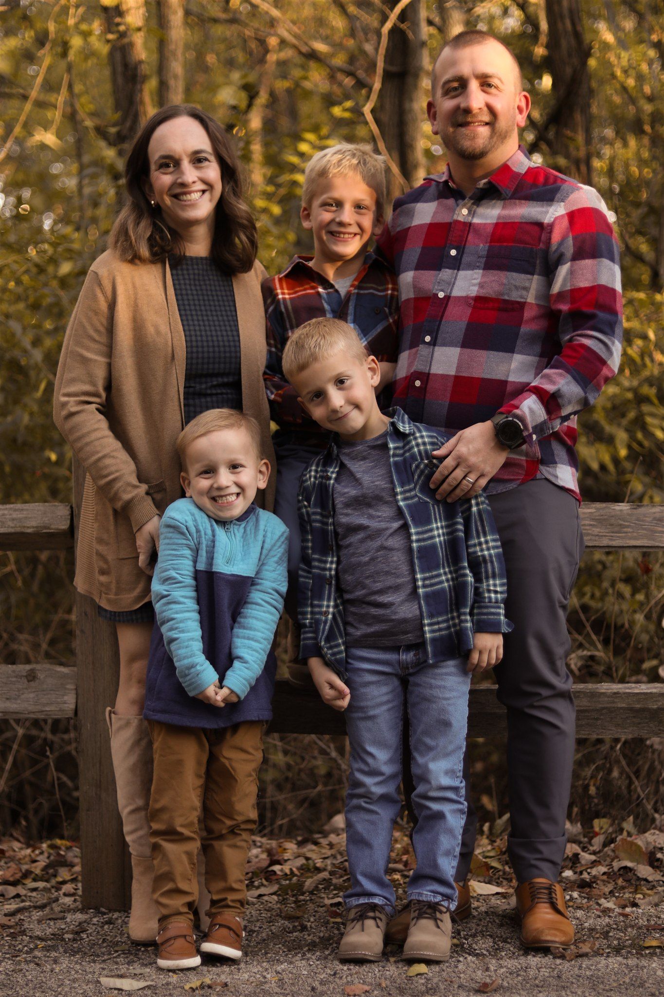 Family wearing coordinated casual outfits poses together on wooden bench in autumn forest setting.