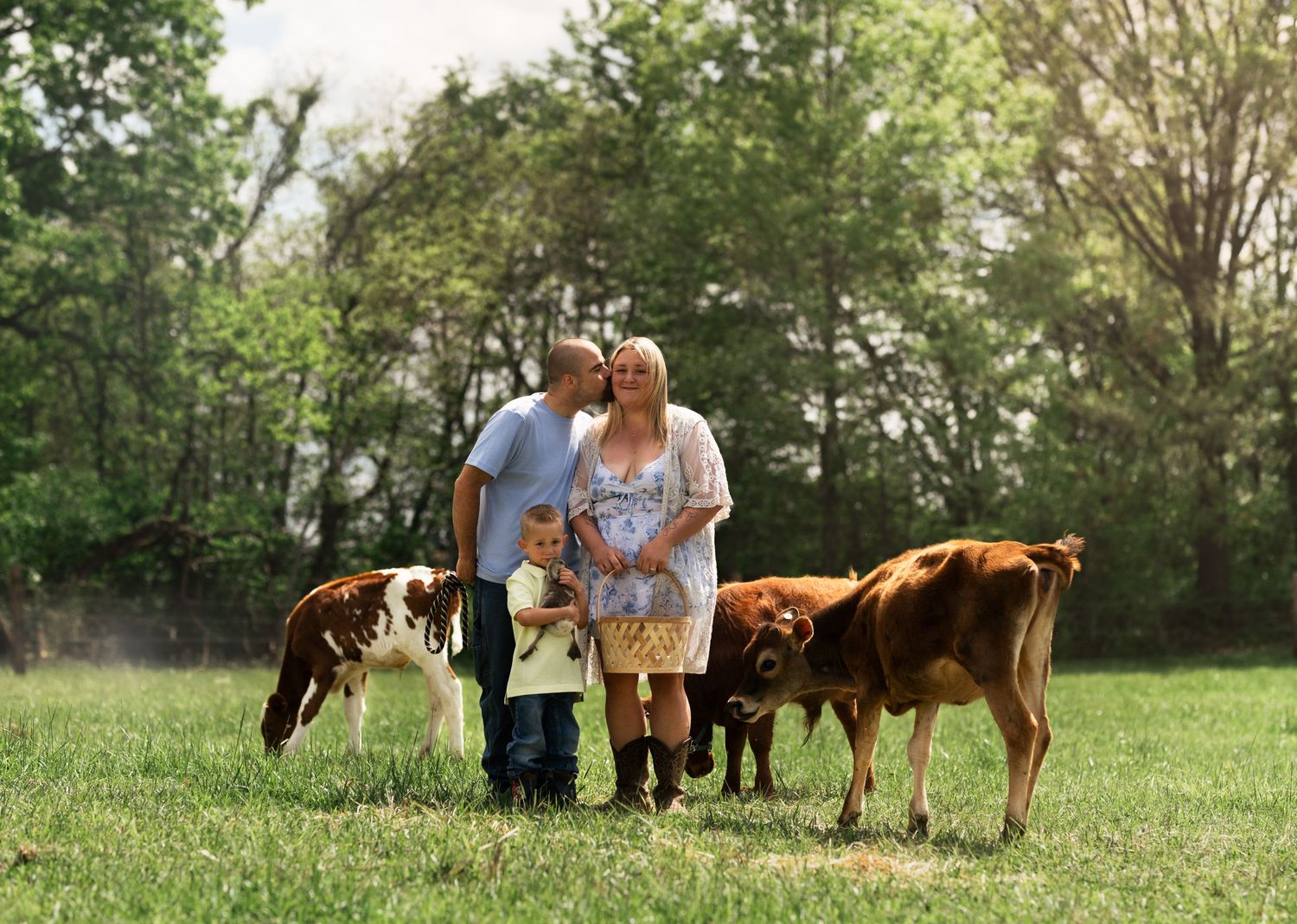 Family with young child walking among calves in a lush green pasture, man kissing woman on cheek.