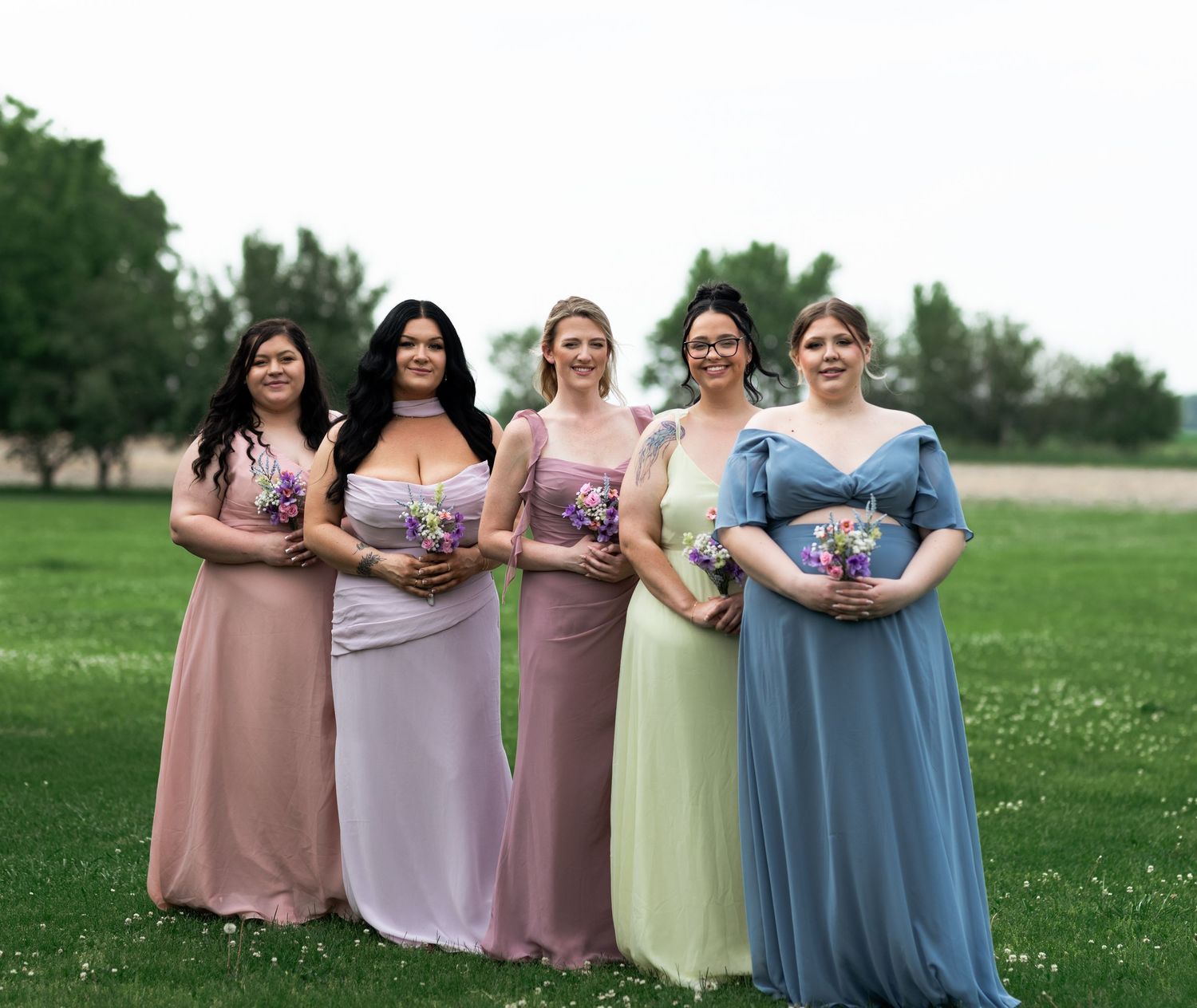 Five bridesmaids in mismatched pastel gowns holding bouquets stand on green grass at an outdoor wedding ceremony.