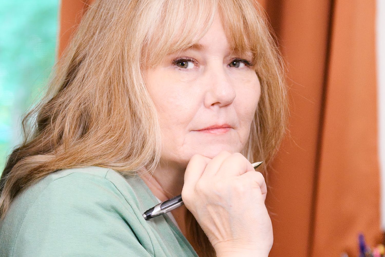 Thoughtful blonde woman holding pen to chin with orange curtains in background.