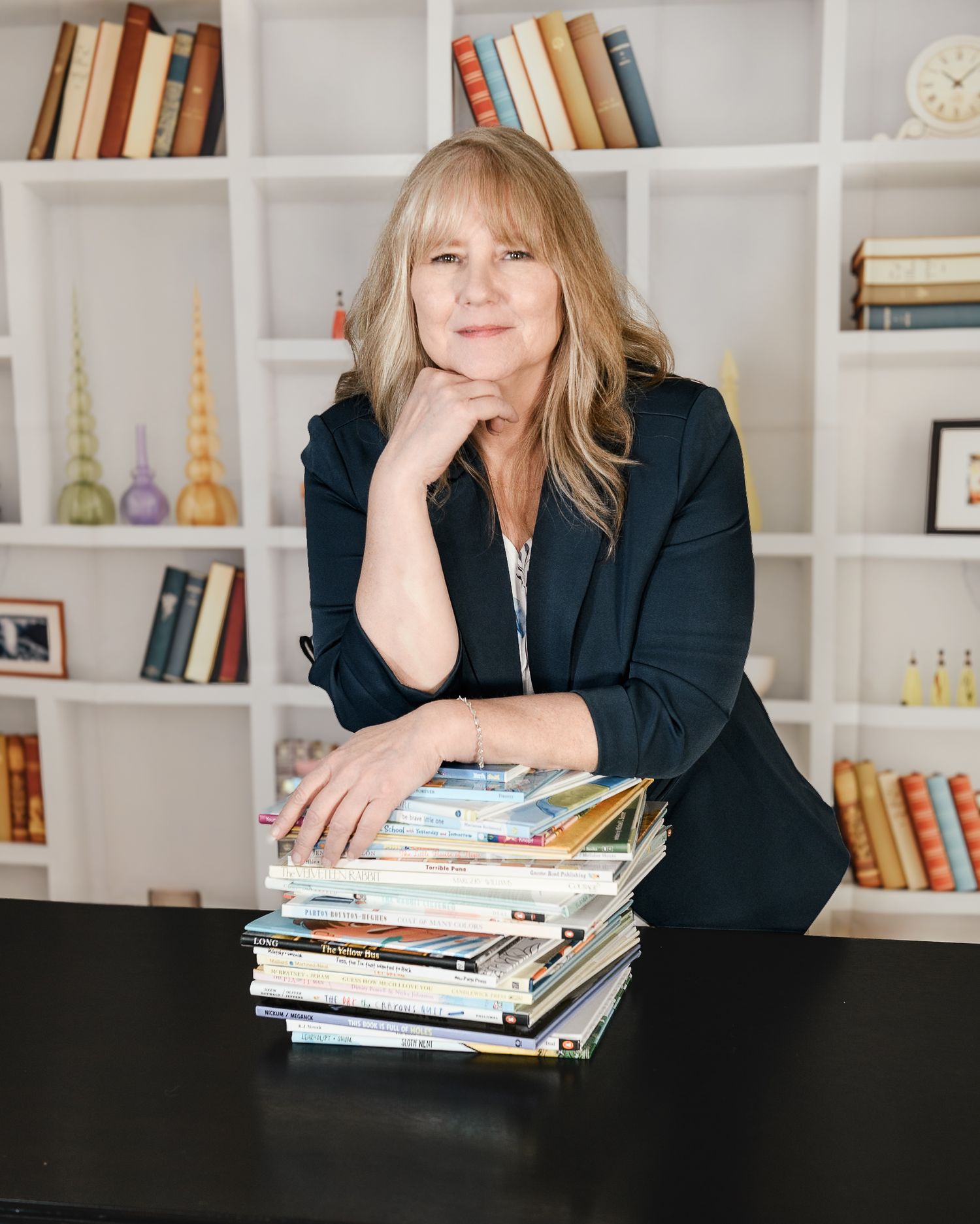 Woman with blonde hair in black blazer posing with stack of children's books in front of white bookshelf.