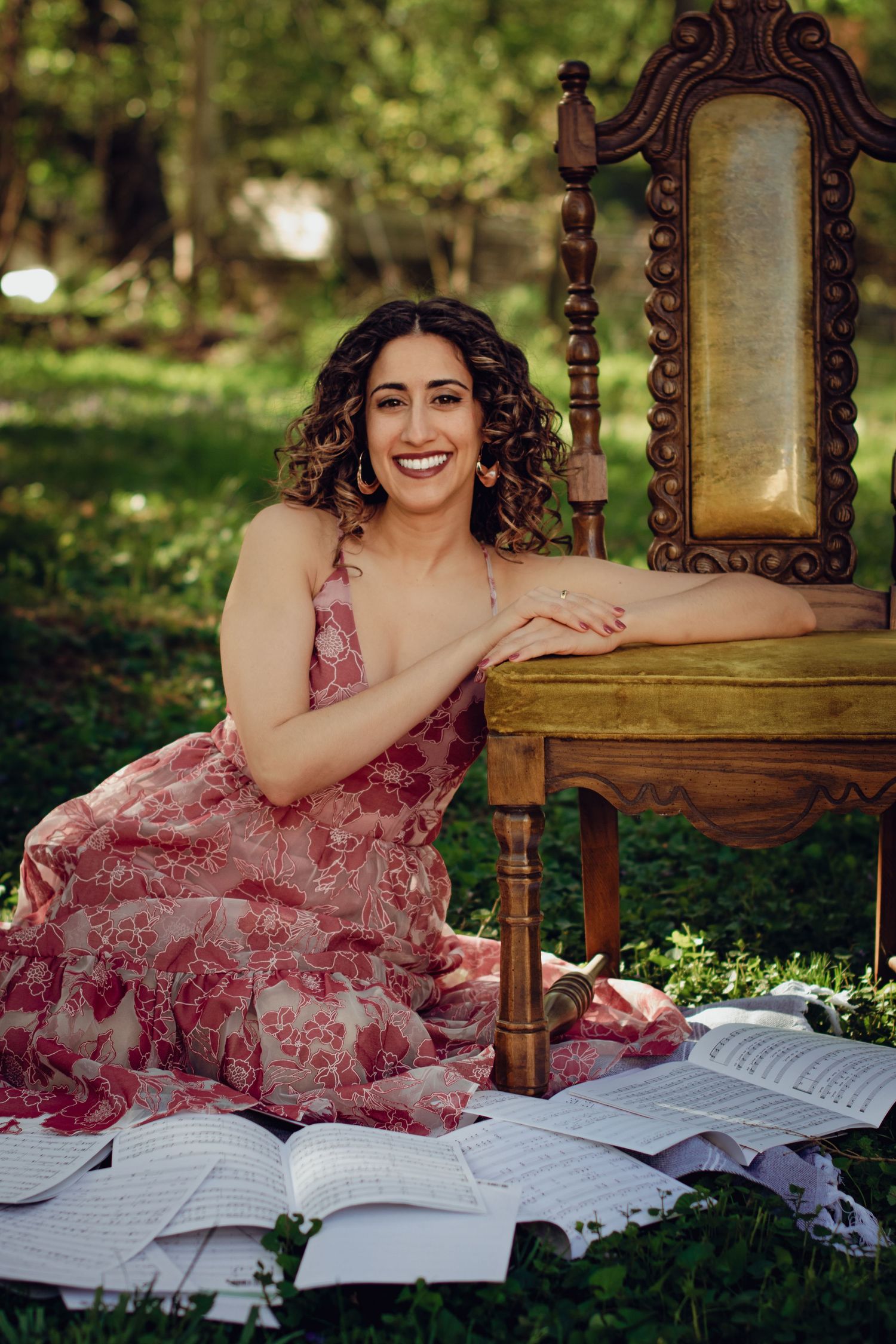 An elegant woman in a floral pink dress in an outdoor garden setting with vintage chair and scattered sheet music. Professional photography.