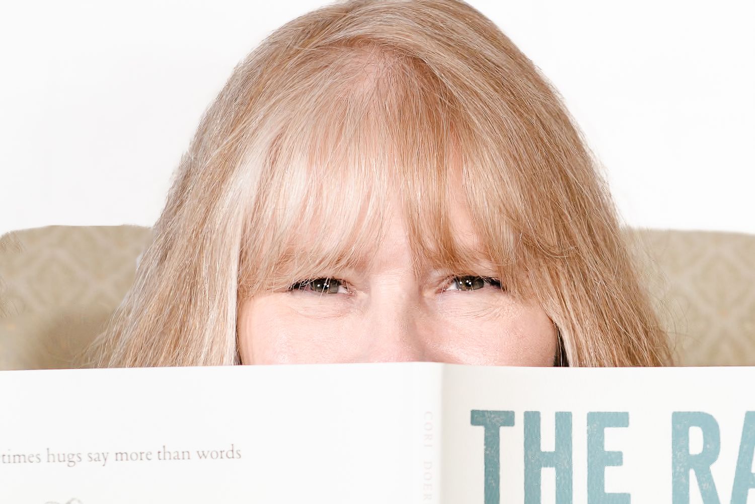 Blonde woman playfully hiding her face behind an open book, peeking over the top with curious eyes.