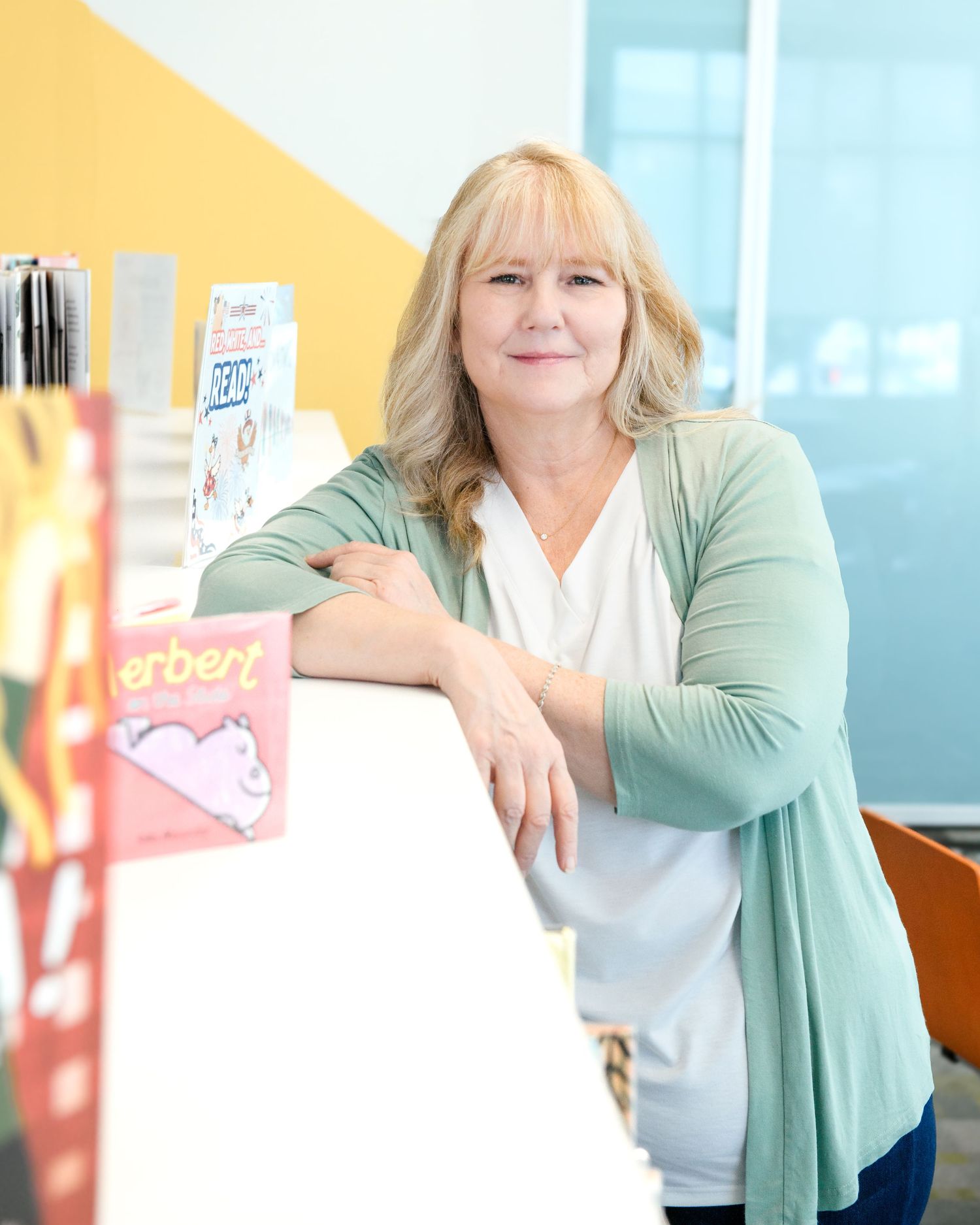 Smiling blonde woman in green cardigan leaning on a bookshelf displaying colorful childrens books.