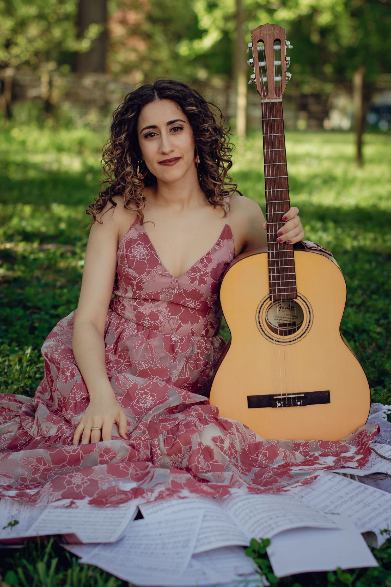 Woman in pink floral dress sitting on grass in park holding acoustic classical guitar with sheet music nearby.