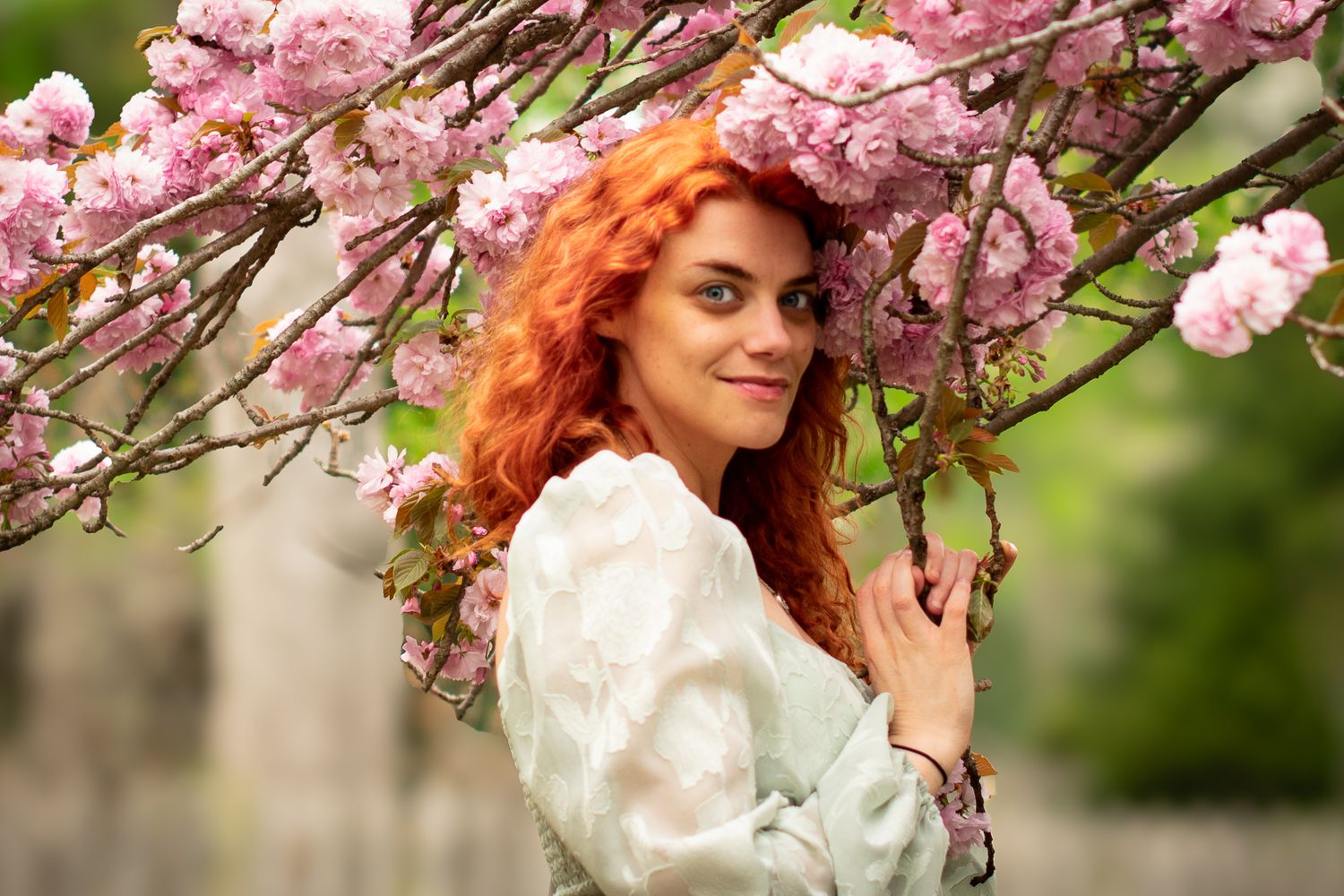 A professional portrait of a woman under pink cherry blossoms wearing mint top with red curly hair against a soft bokeh background.