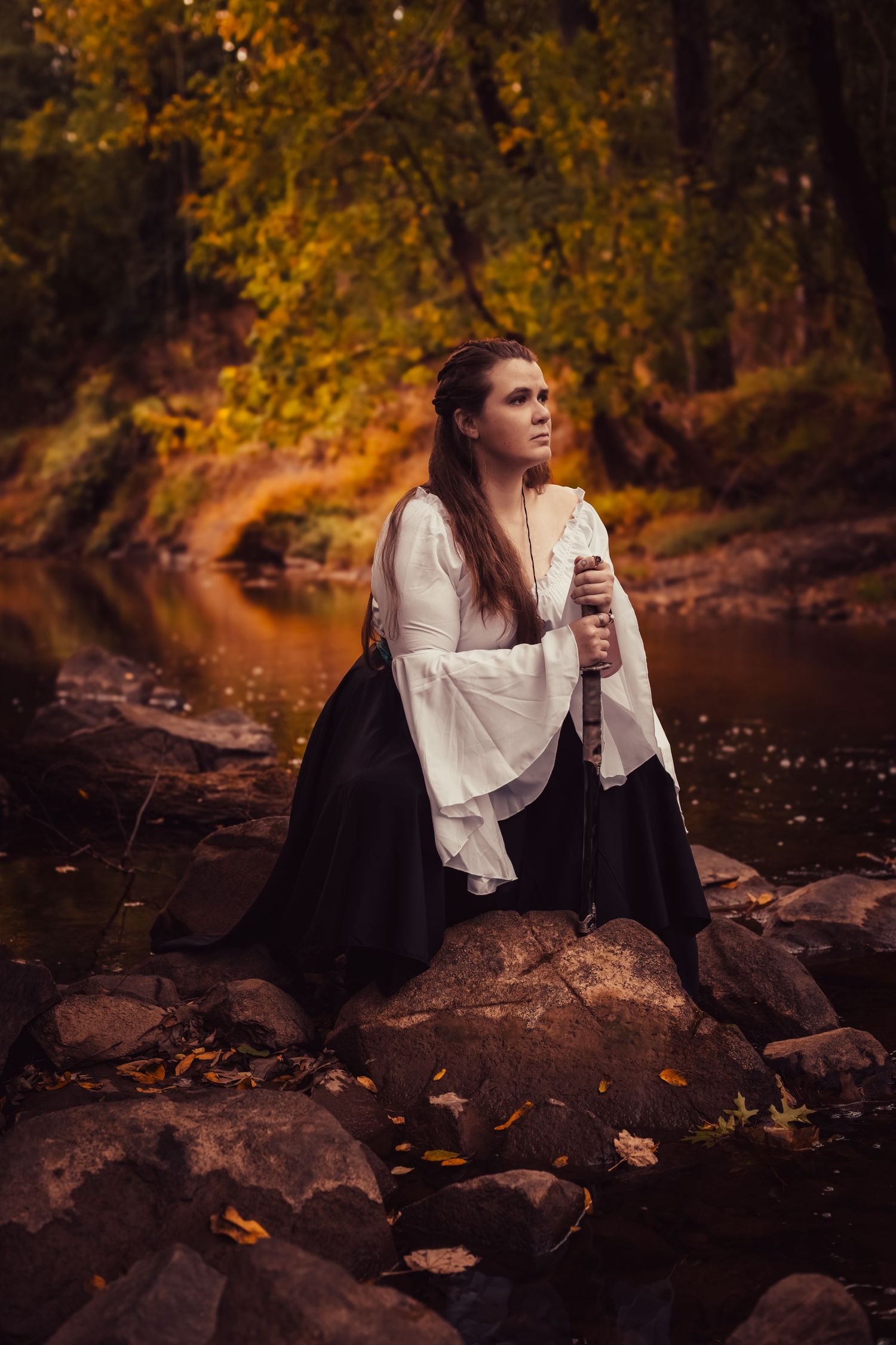 A figure in a white blouse sits thoughtfully on rocks by a stream during autumn with golden foliage in the background.