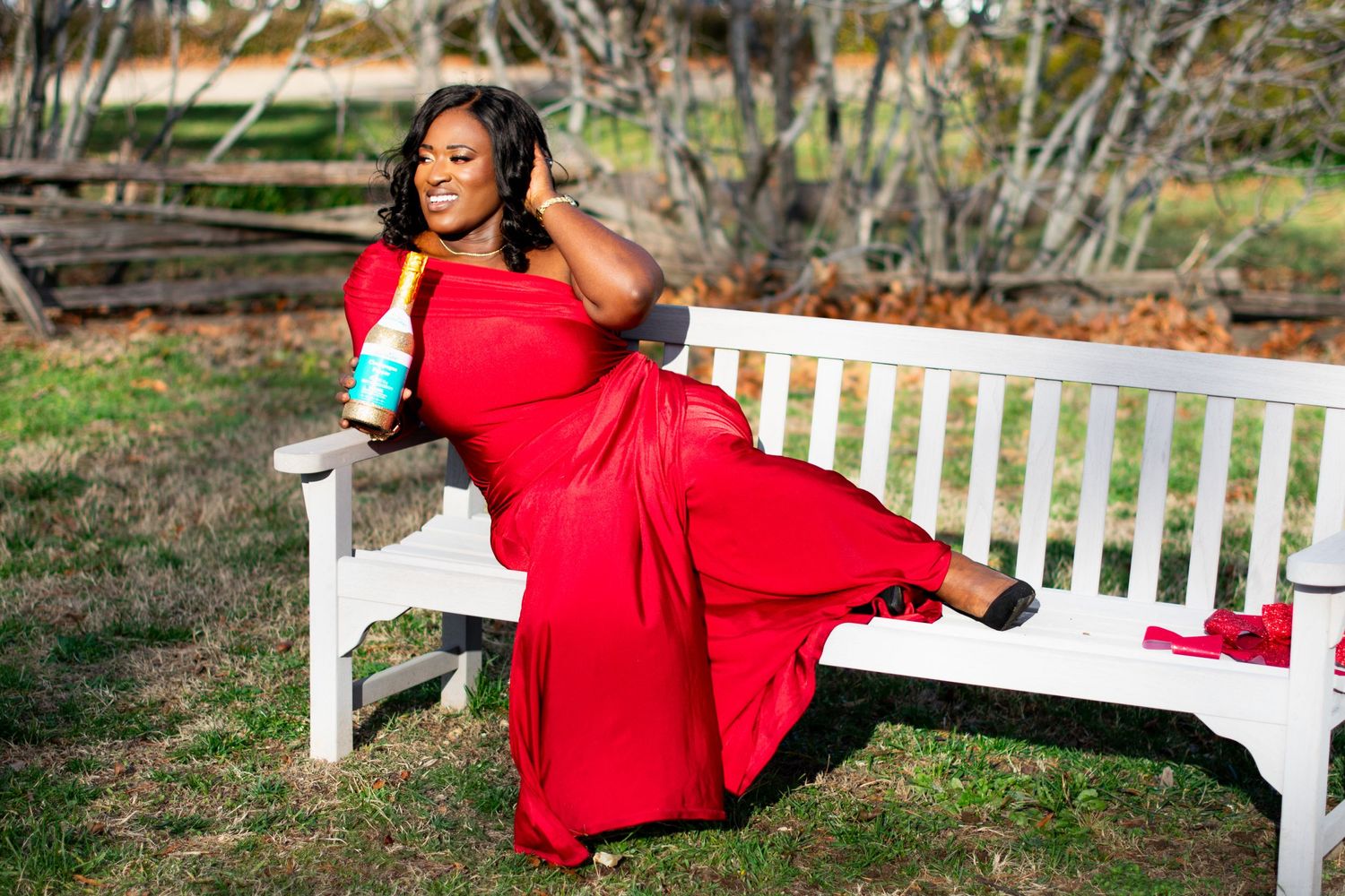 Woman in red one-shoulder dress relaxing on white bench with champagne bottle. Elegant outdoor portrait in natural setting.