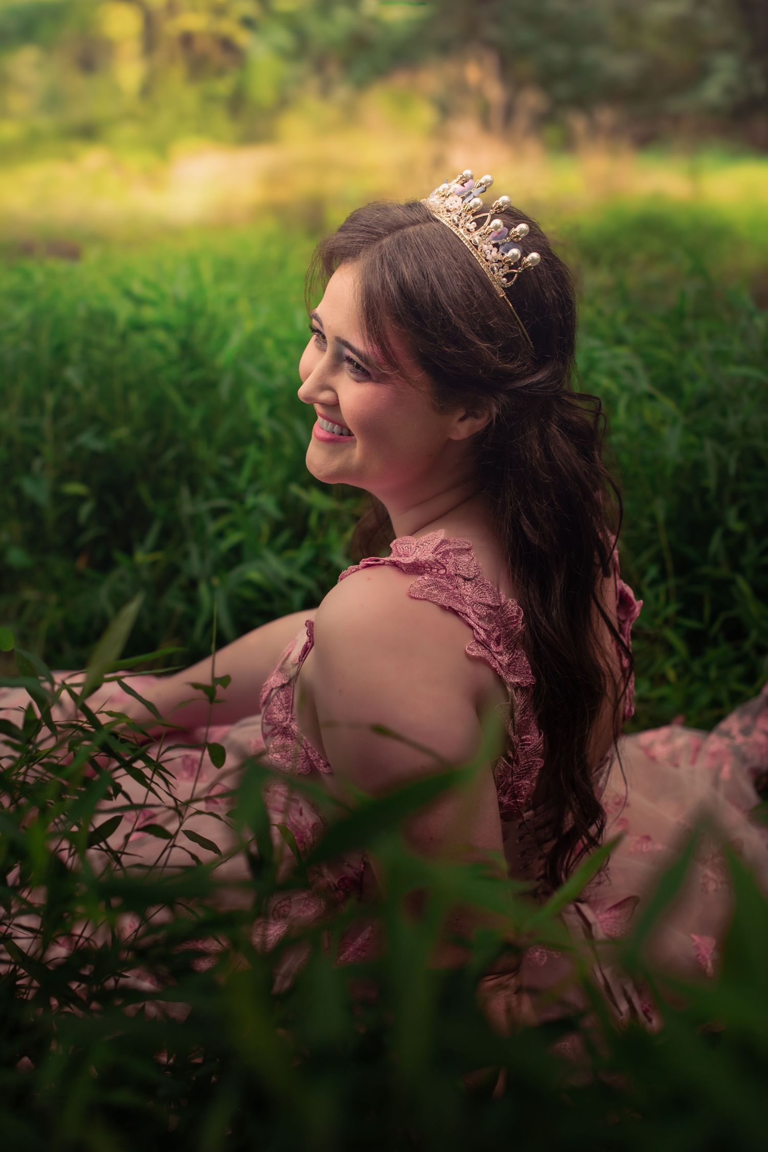 Side profile portrait in pink dress and tiara amid lush garden foliage.