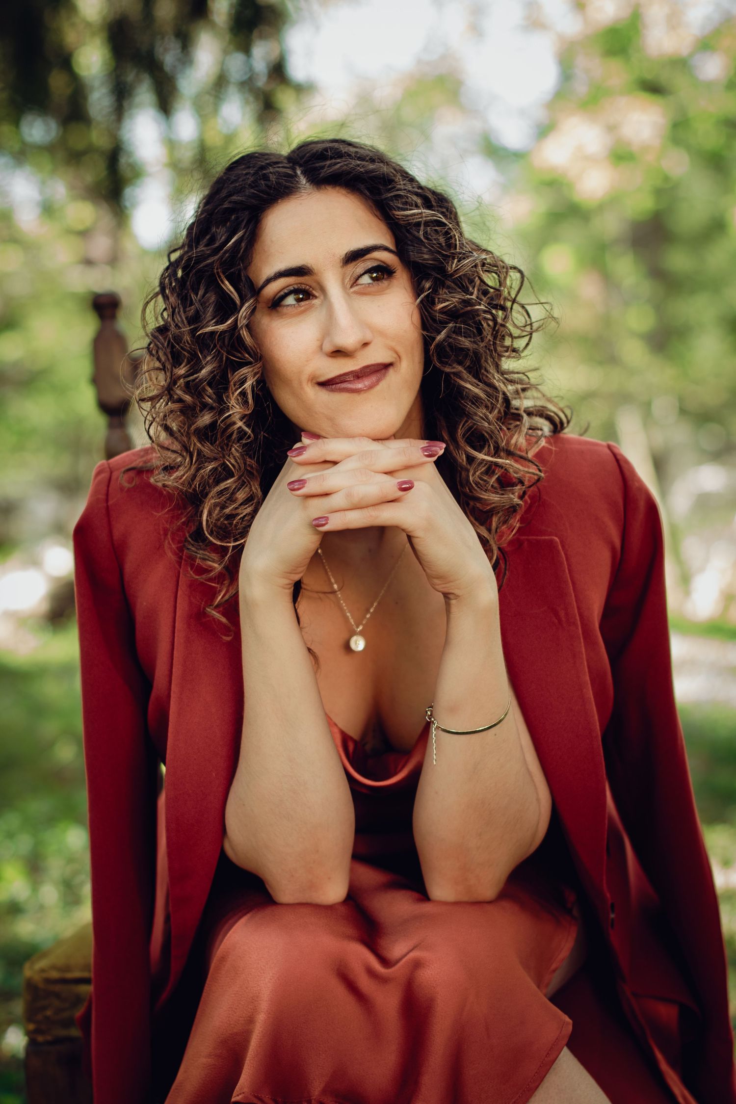 A professional portrait of woman outdoors featuring curly hair, red blazer, and delicate necklace against a soft natural background.