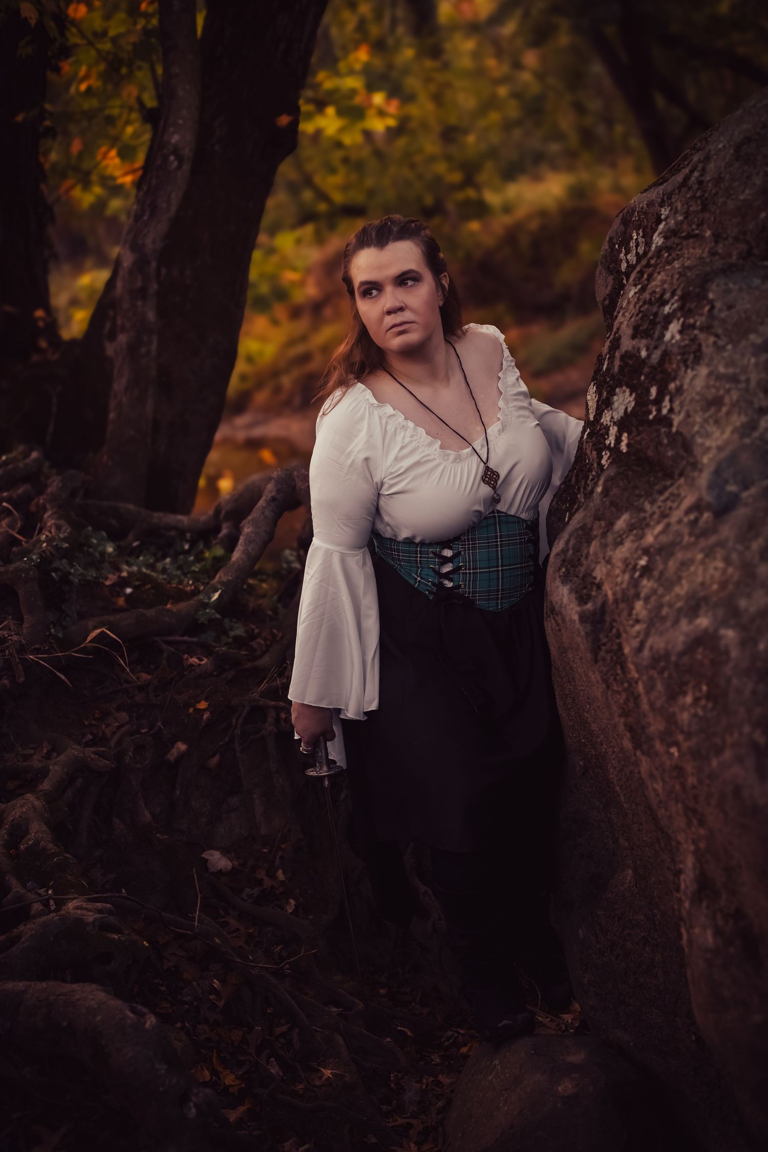 Woman in white blouse and teal corset poses among autumn forest rocks in fantasy-inspired costume.