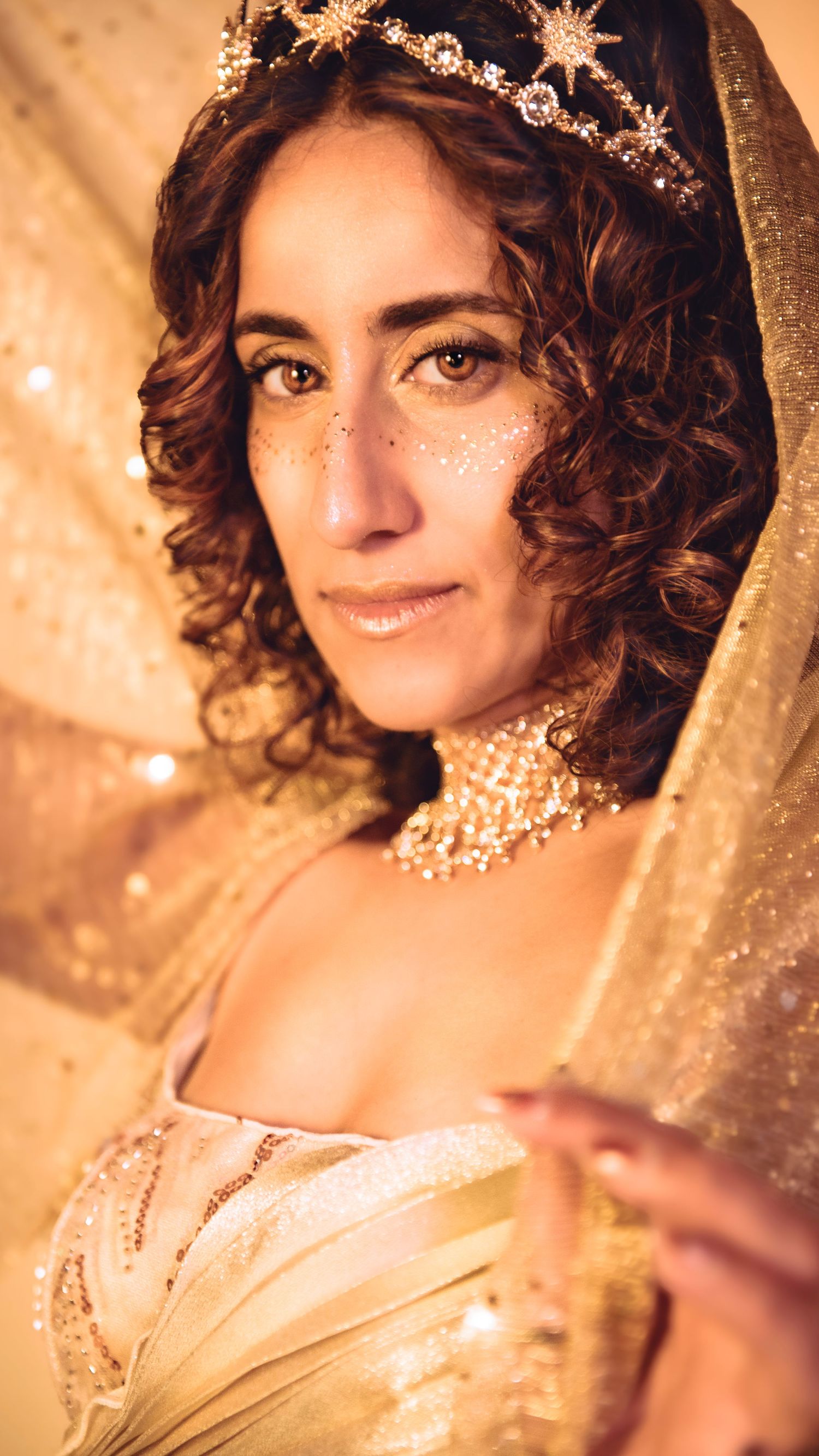 A woman in a gold bridal look with a sparkling tiara, glitter freckles, and a shimmering veil and choker necklace.