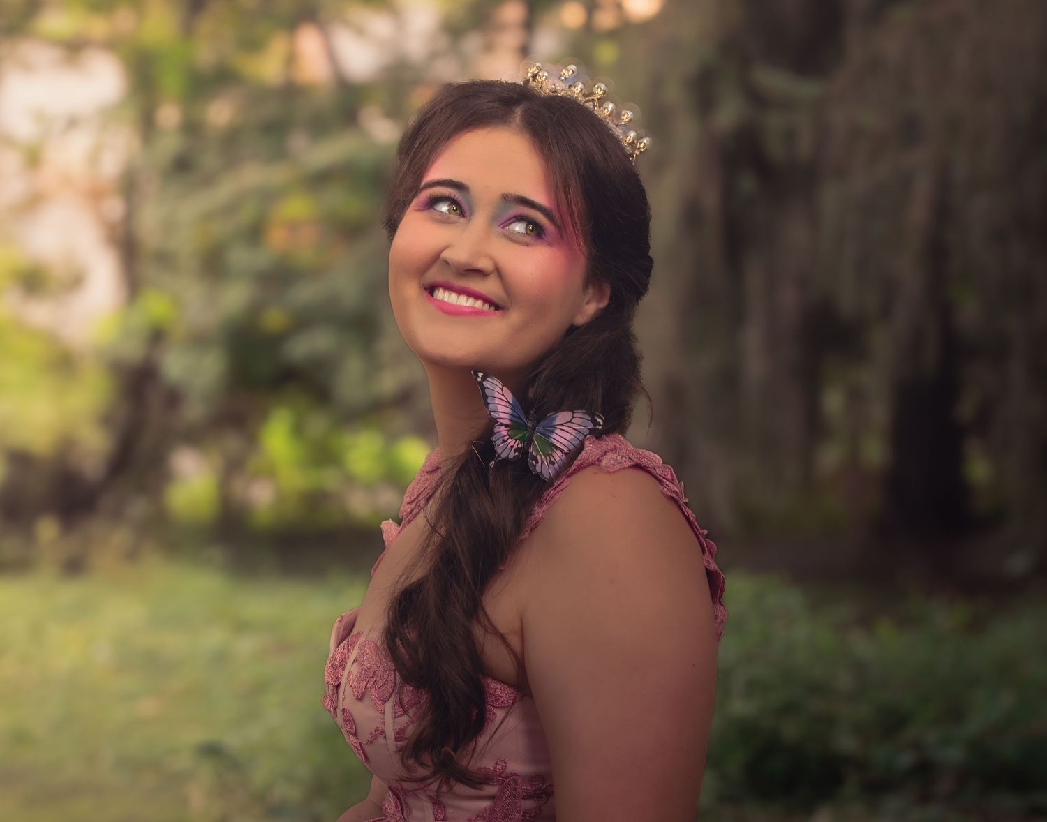Figure wearing tiara and pink dress smiles while looking up in forest setting.