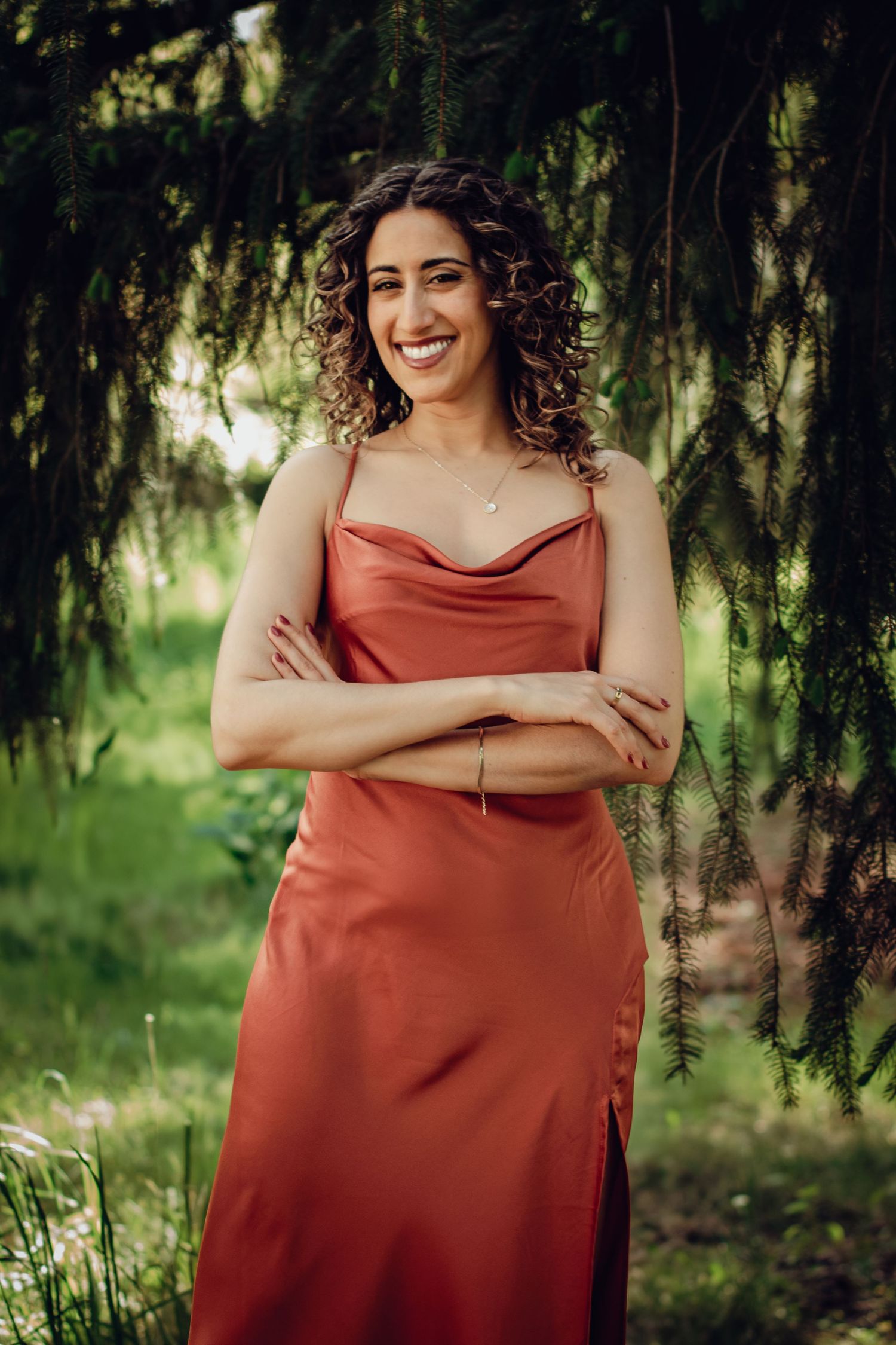 An empowered woman wearing a rust-colored satin dress poses with crossed arms in a garden setting with lush greenery. Professional portrait