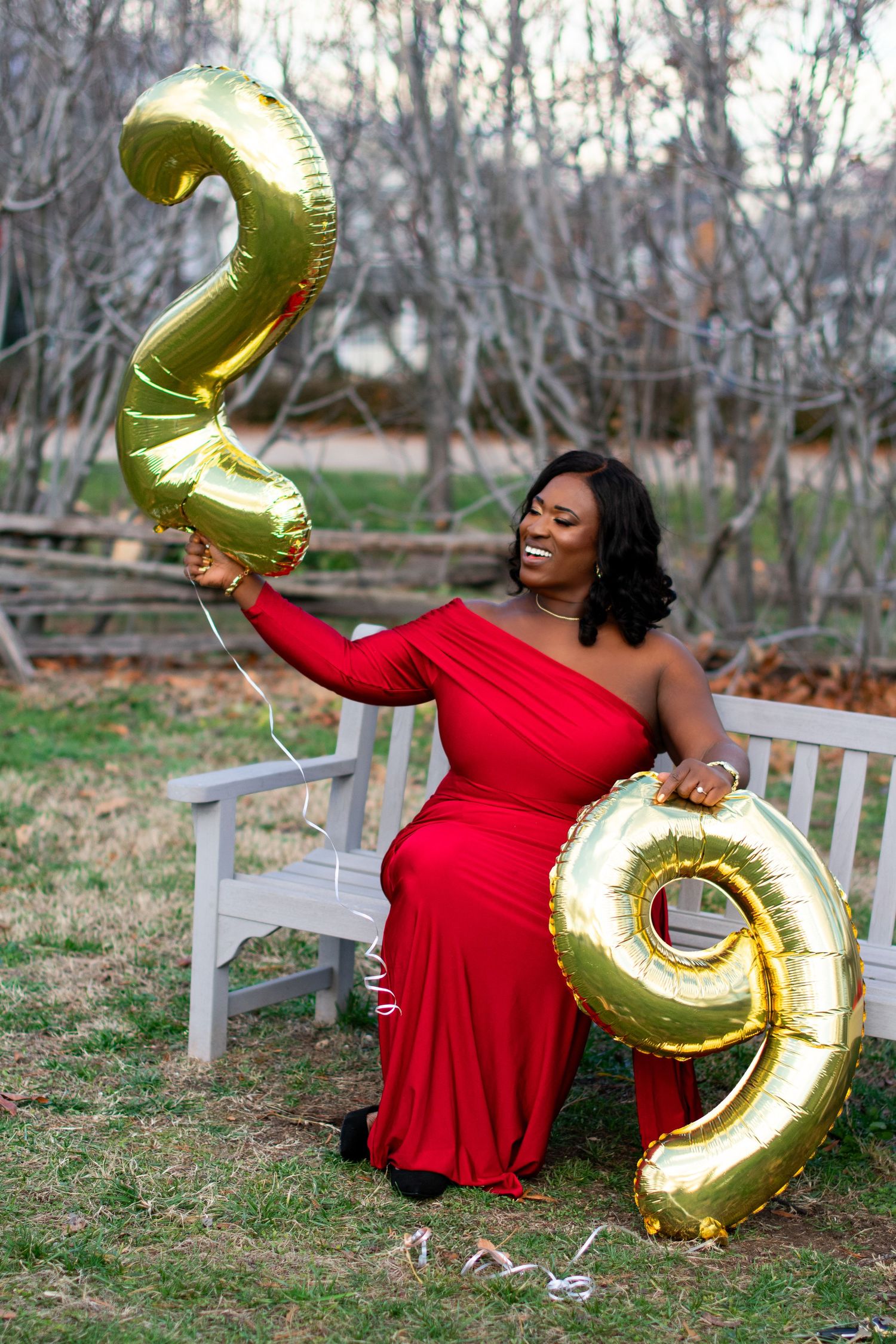 Birthday celebration portrait with woman in red dress holding gold number '29' balloons. Outdoor photography with natural backdrop.