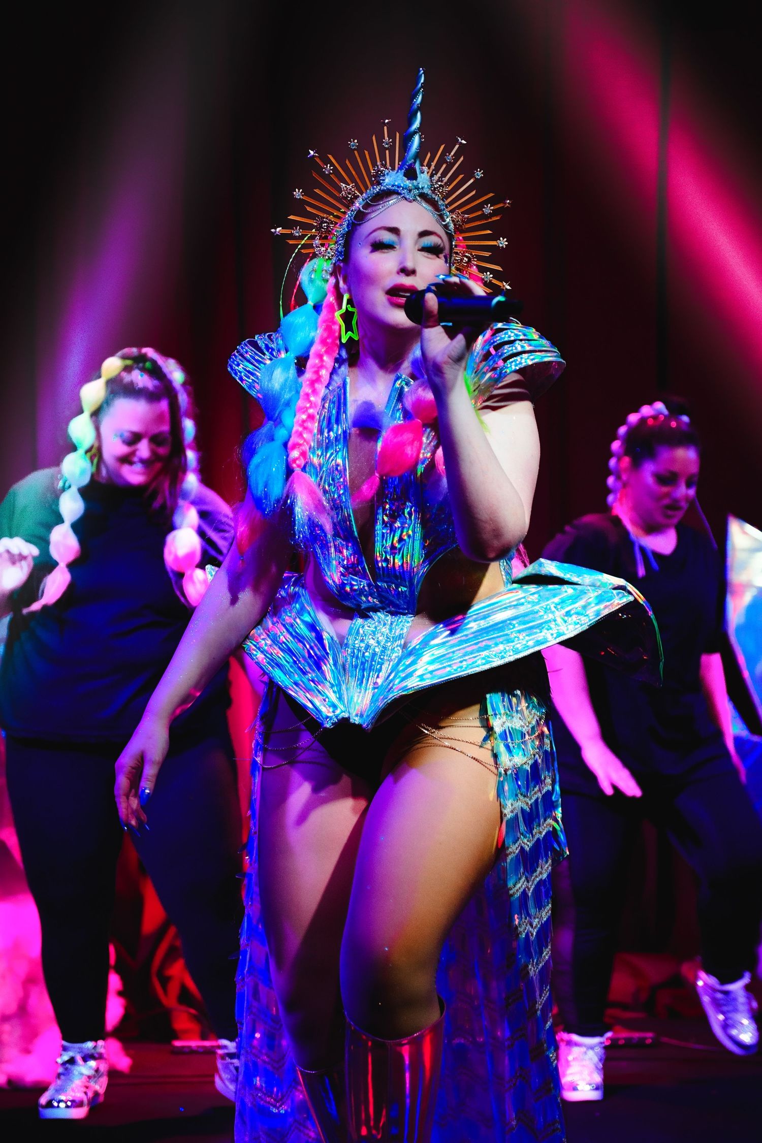 Female performer in blue sequin costume and crown headpiece singing on stage with dancers.