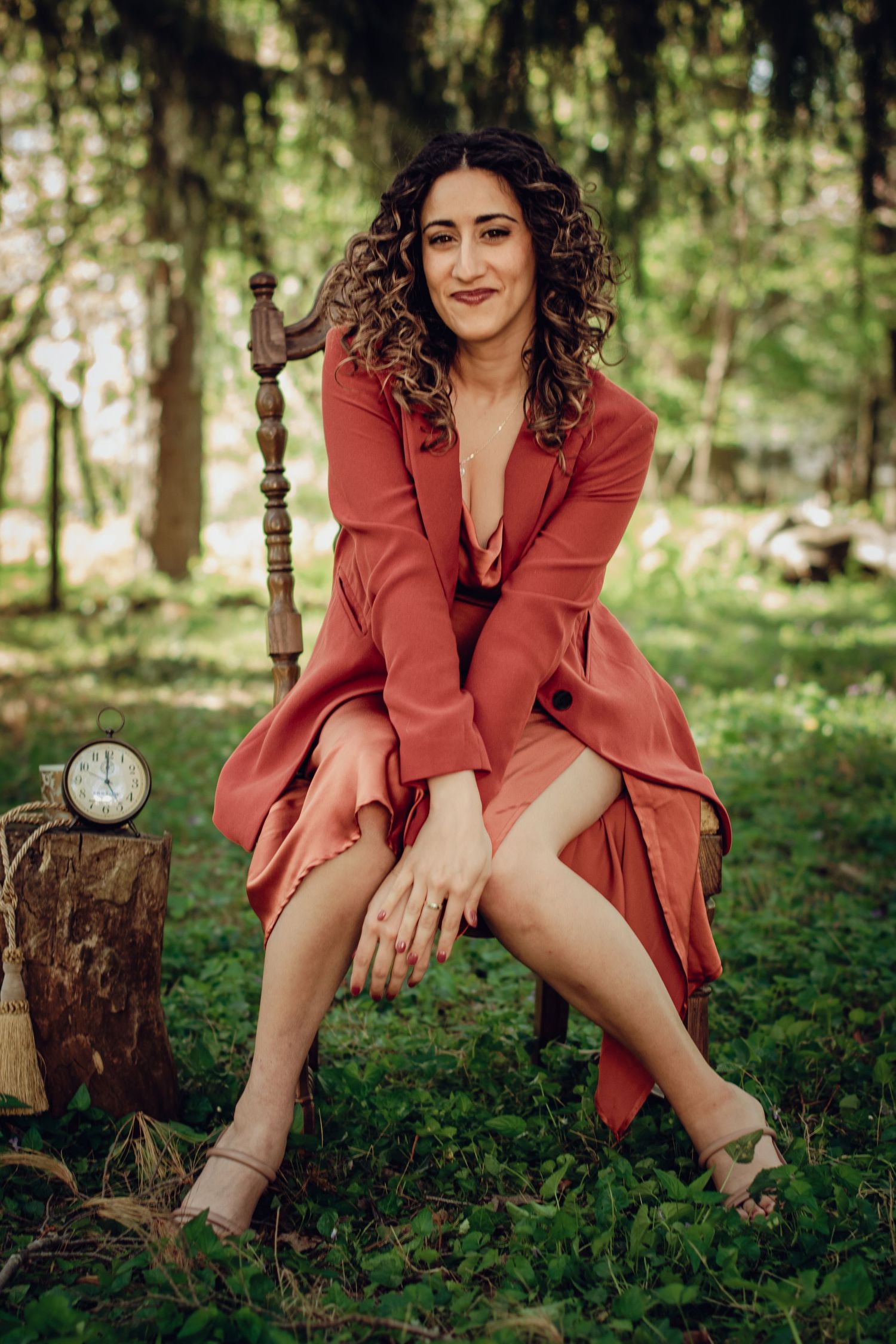 Professional woman in rust-colored dress poses outdoors sitting next to a vintage clock in a dreamy forest setting. Professional portrait.