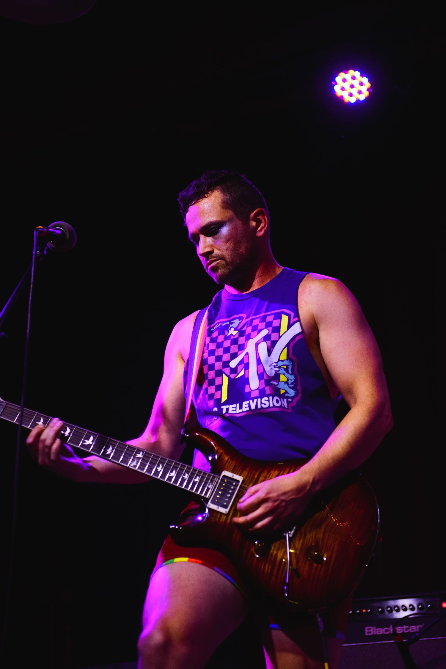 Male guitarist performing on stage wearing MTV tank top under purple spotlight at live music event.