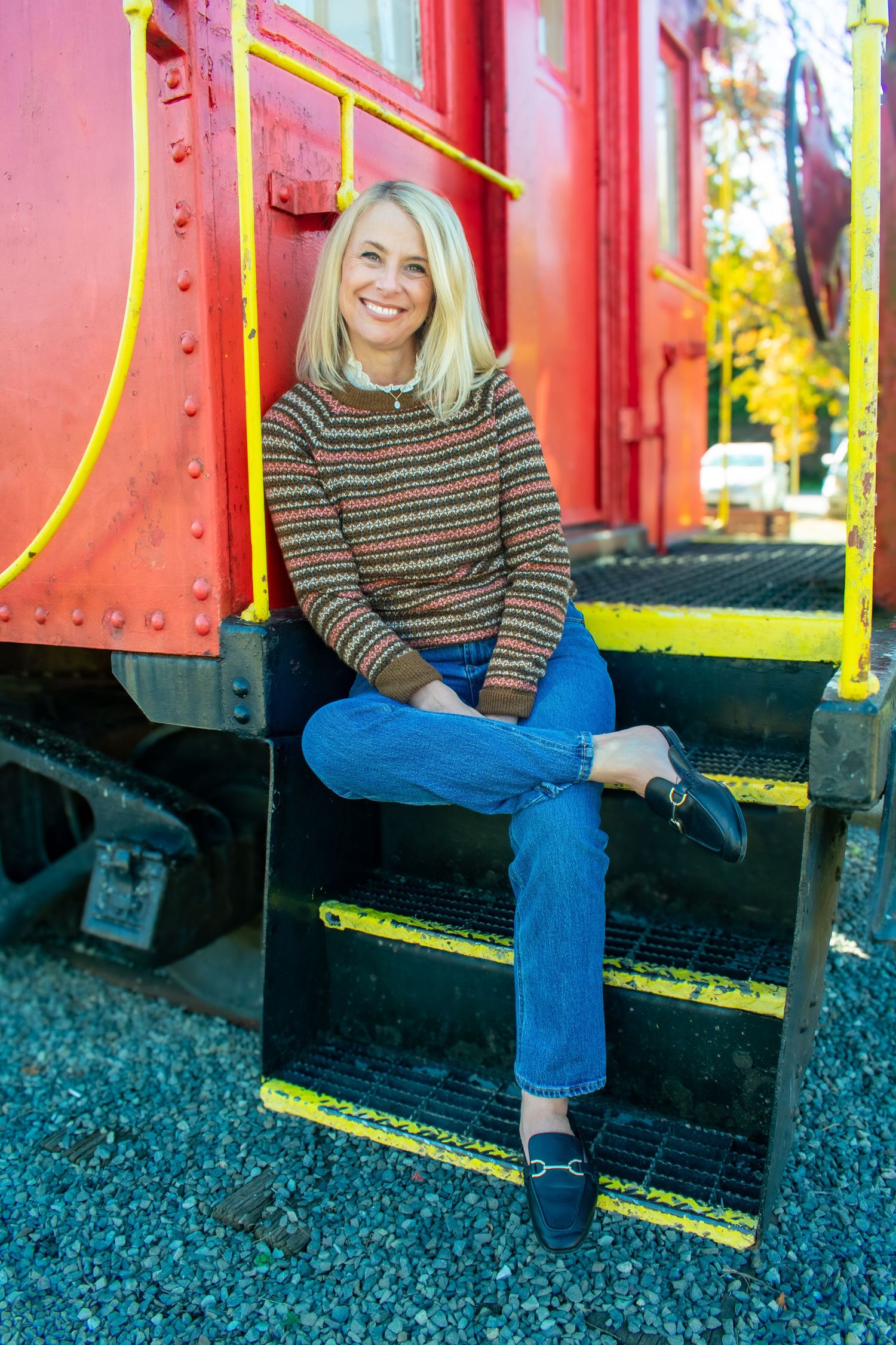 Person in striped sweater and blue jeans sitting casually on red train car steps with yellow railings.