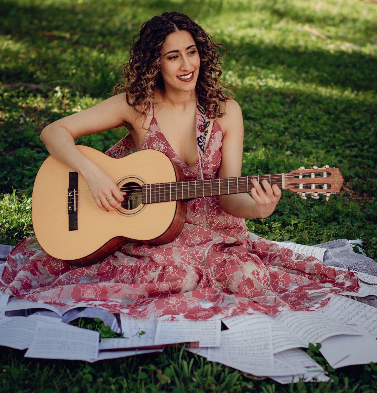 Woman playing acoustic guitar while sitting on blanket with sheet music scattered on sunny grass lawn. Professional portrait photography.