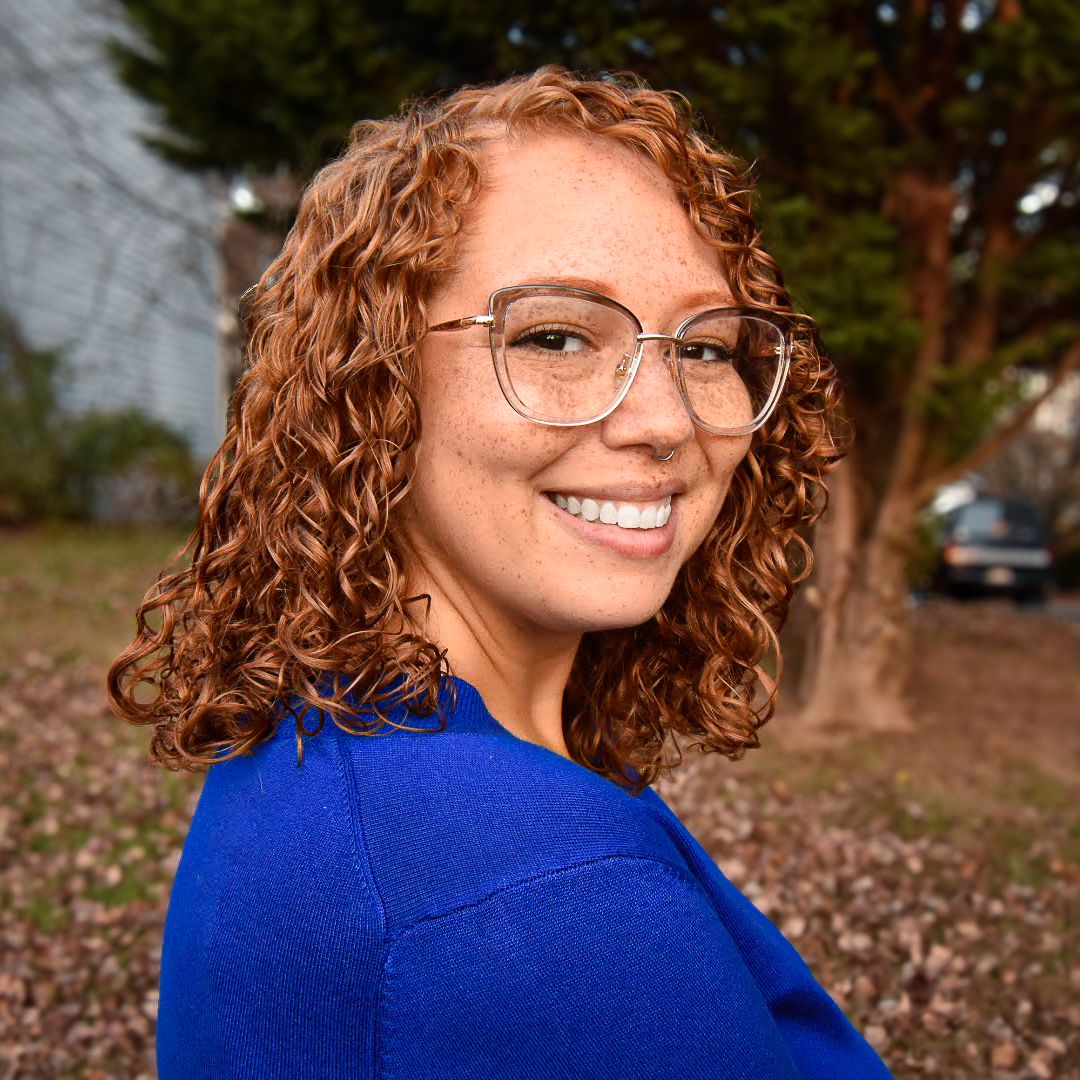 Outdoor portrait wearing glasses and blue sweater with autumn trees in background.