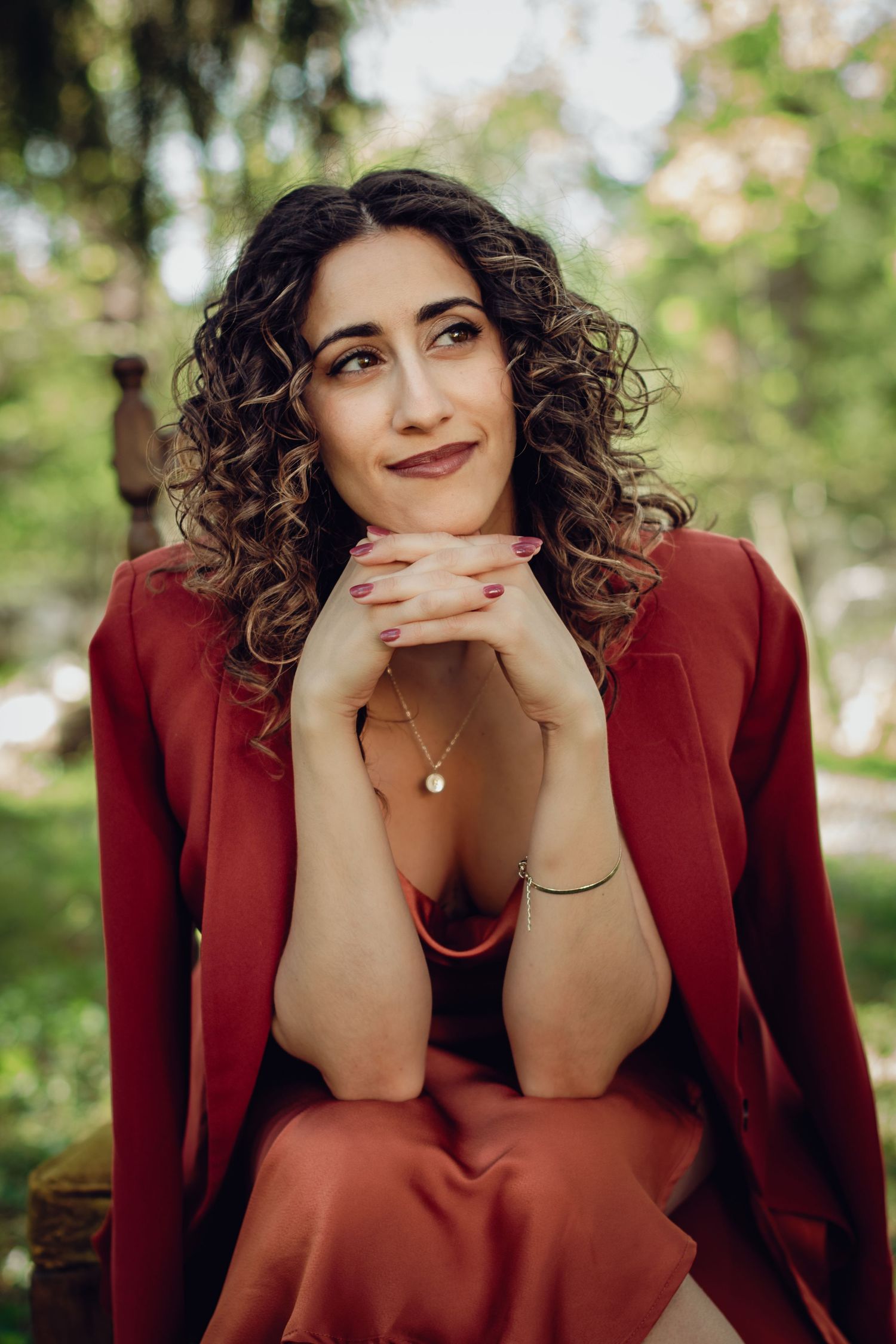 Outdoor portrait of someone in a burgundy blazer with curly hair seated in a natural setting.