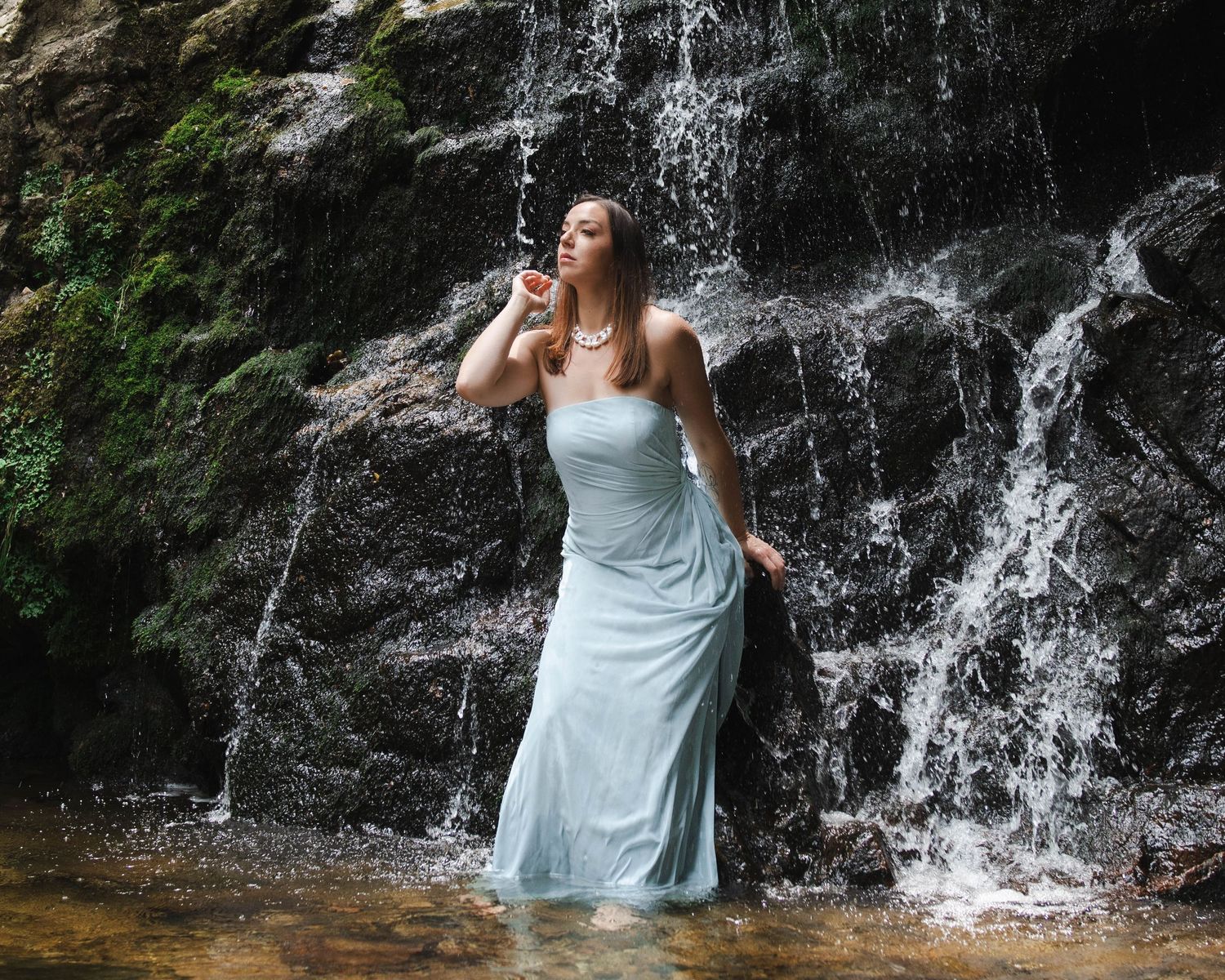 Woman in white dress standing before a cascading waterfall, water flowing around her in a serene natural setting.