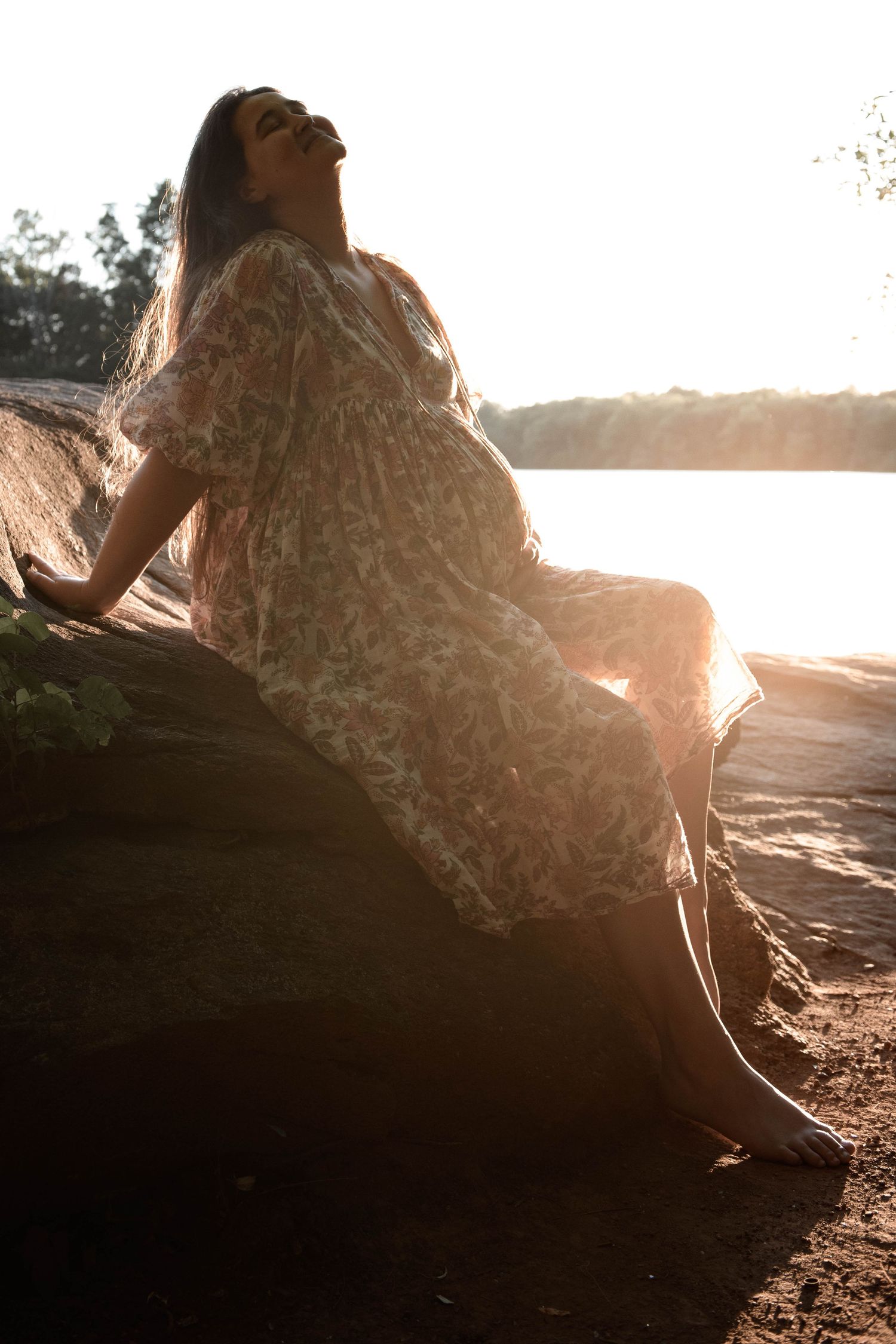 Pregnant woman in floral dress sitting on a log by a lake, bathed in golden sunset light.