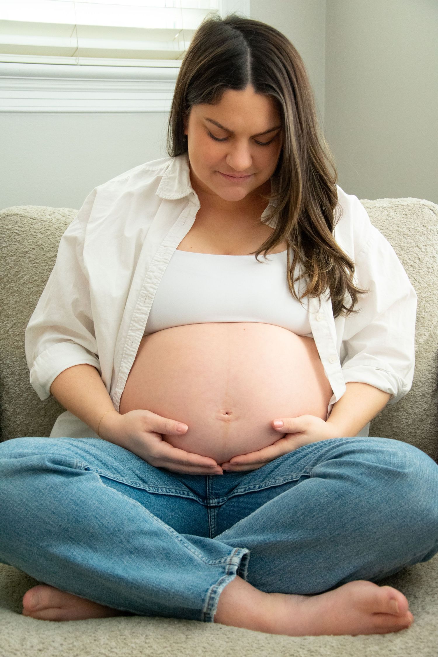 Expectant mother in white shirt and jeans sits cross-legged on couch while cradling pregnant belly. Professional maternity photography