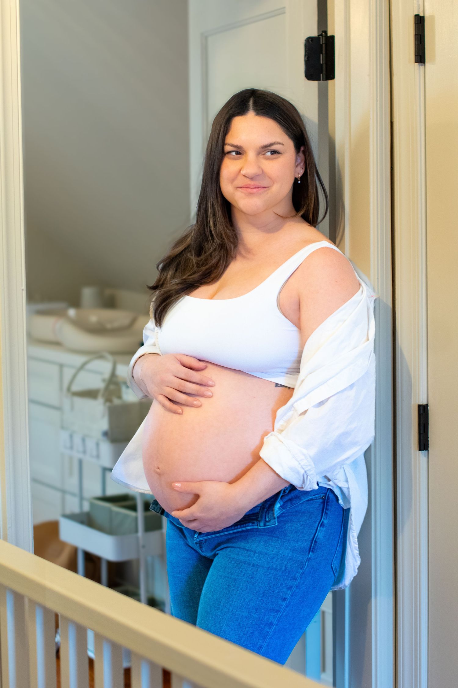 Woman in casual attire leaning against doorframe, posing for maternity photo in a modern home interior with white walls.