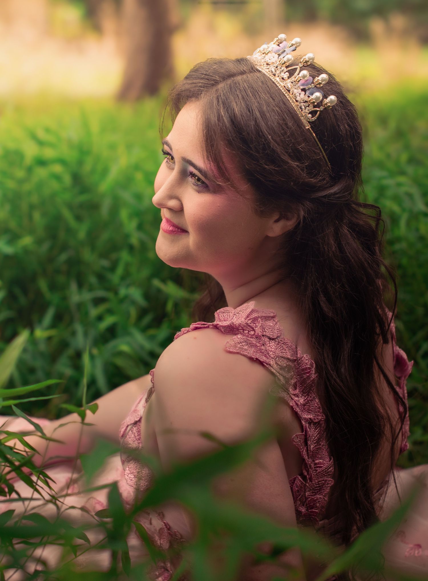 A woman in a mauve lace dress poses outdoors in a dreamy natural setting with soft bokeh effect and green foliage.