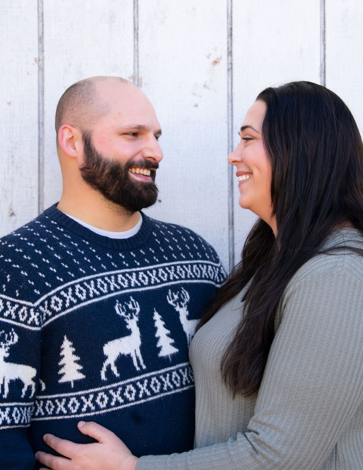 Happy couple in holiday sweaters smile at each other against white wooden background.