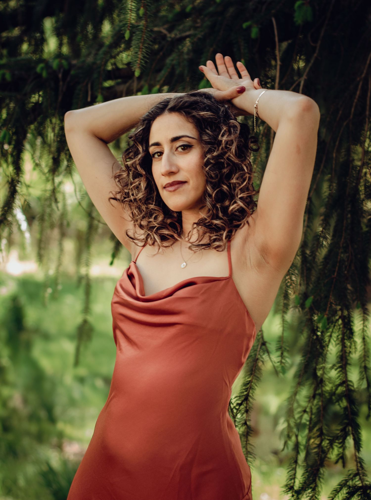 Woman in rustic red dress poses with raised arms against greenery and natural lighting. Professional empowerment portrait photography.
