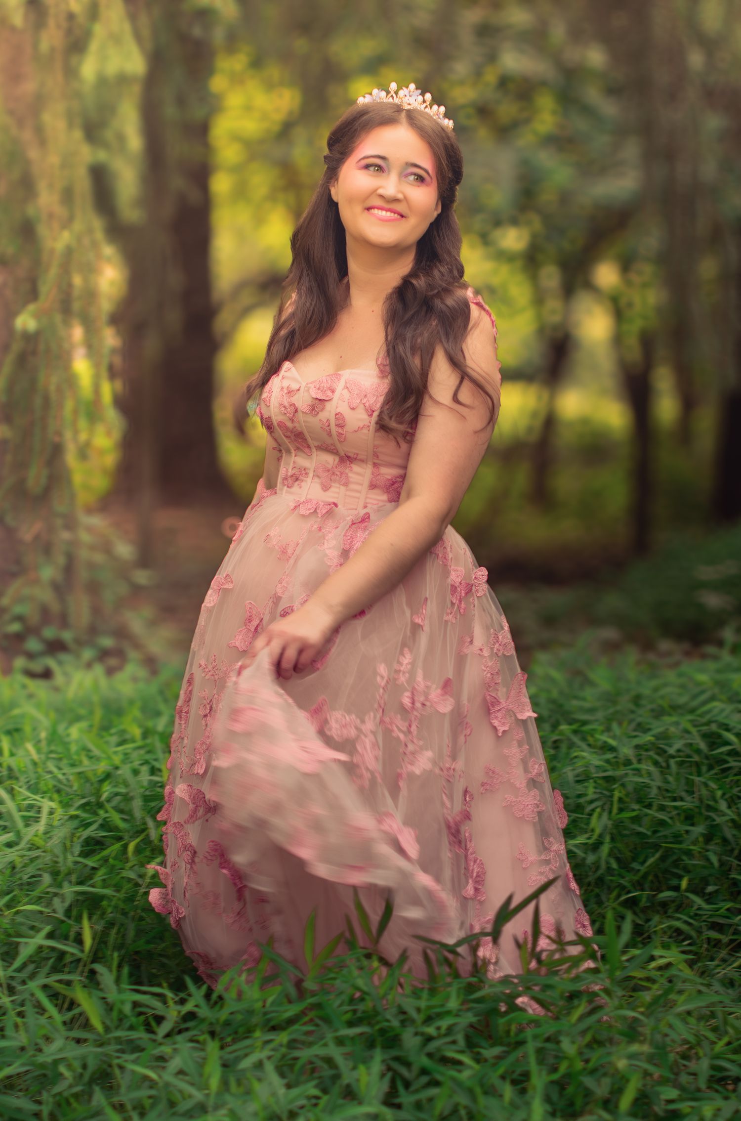 Full-length portrait in pink embellished gown and tiara standing in wooded setting.