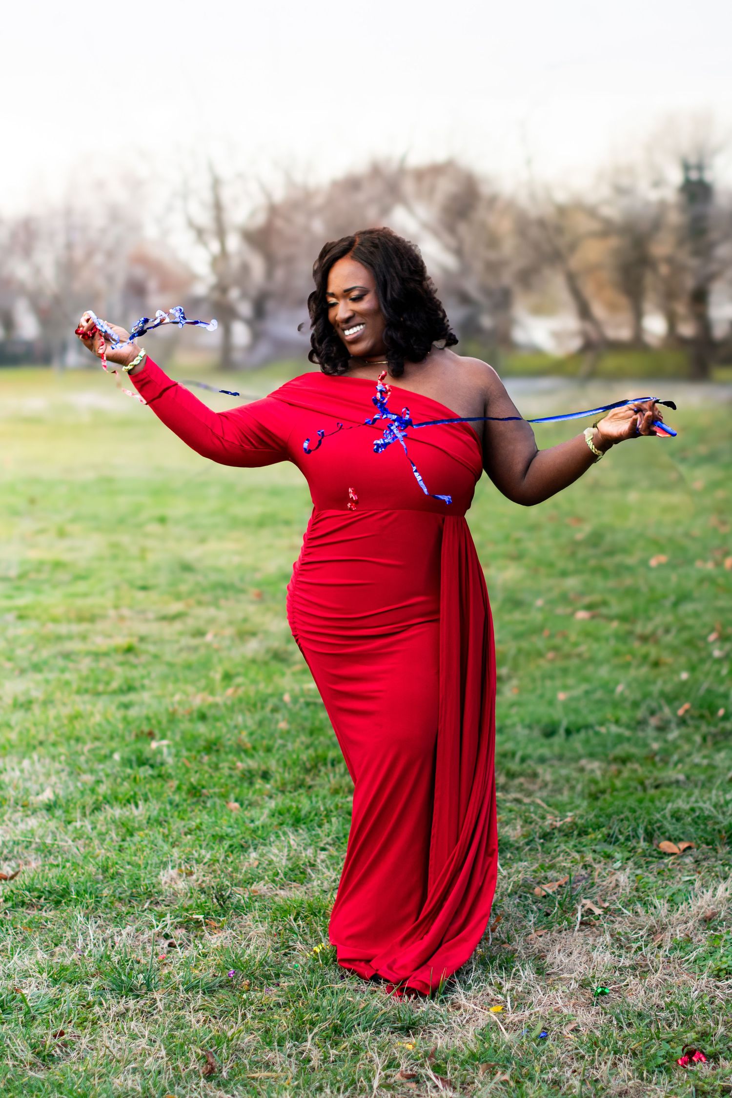 Woman in vibrant red gown celebrating with party streamers in outdoor park setting. Joyful portrait with natural background.