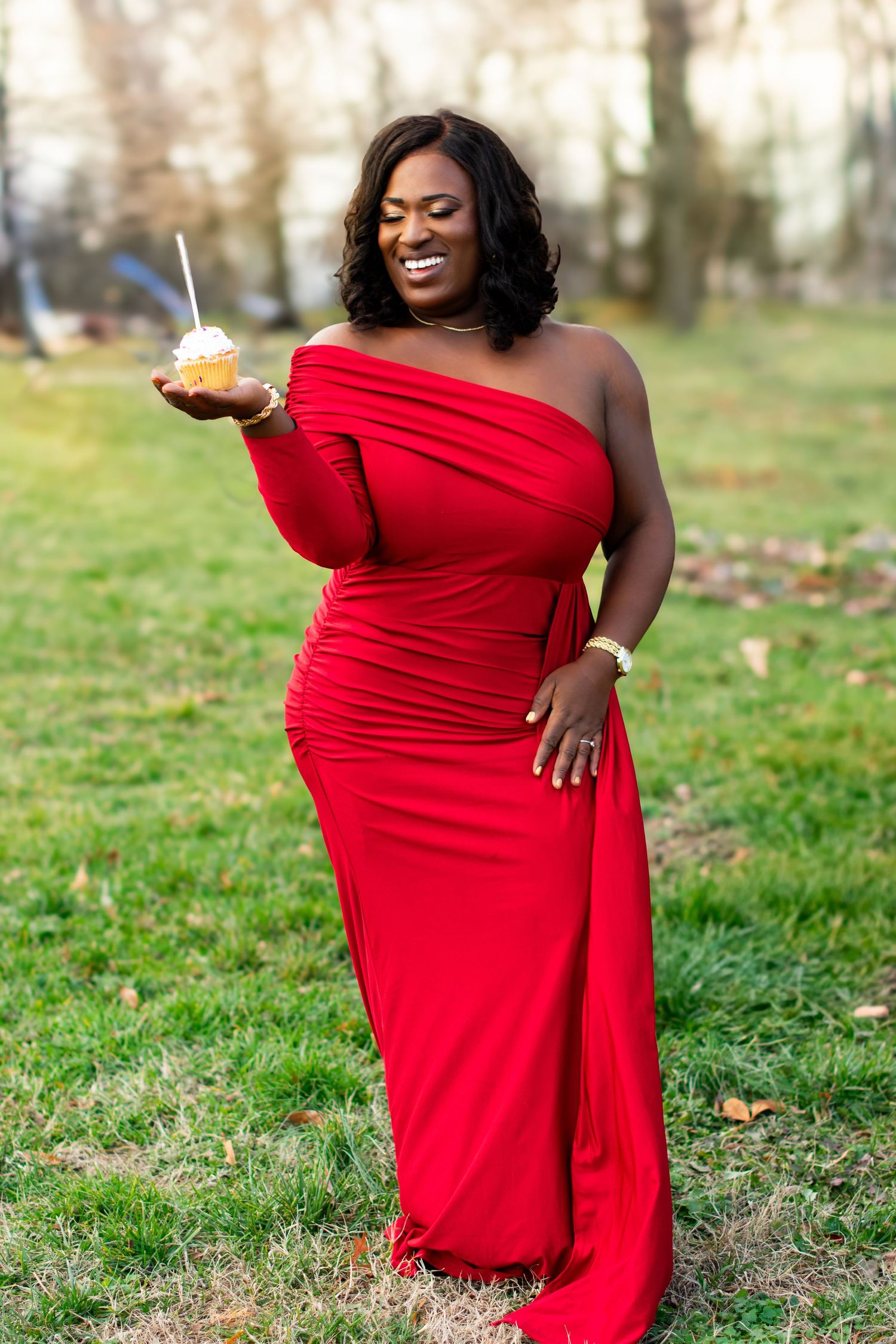 Elegant woman in red one-shoulder dress holding cupcake. Outdoor portrait with soft natural lighting and blurred nature background