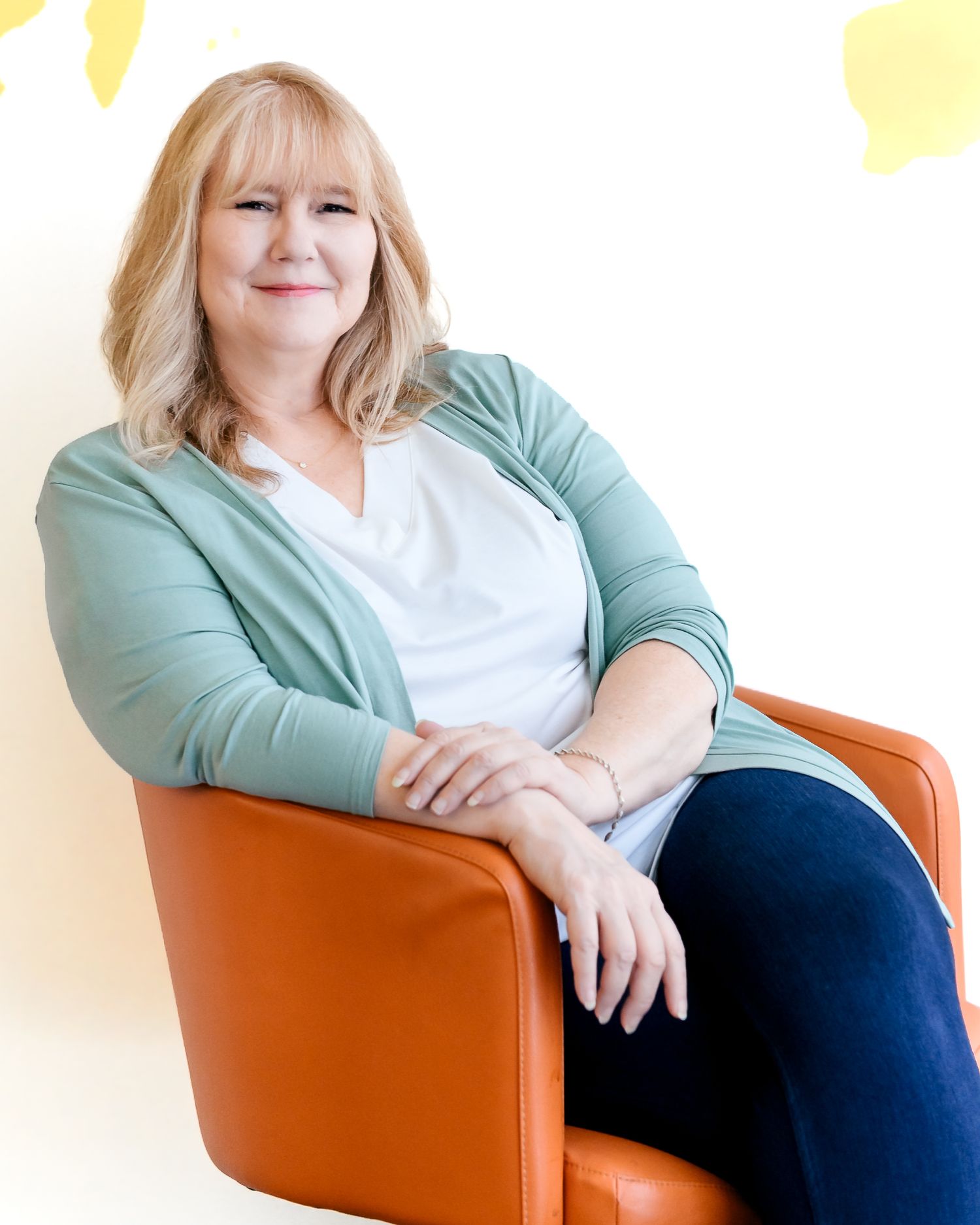 Smiling blonde woman in teal cardigan and white top sits relaxed in an orange chair against a light background.