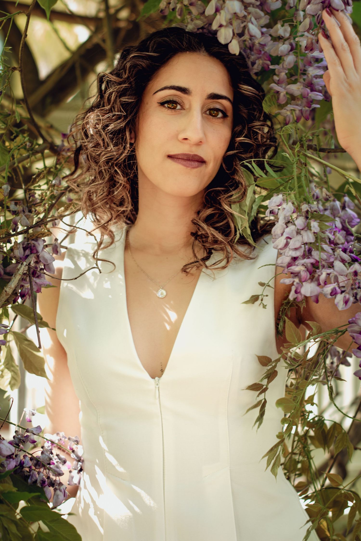 A woman in a white top with curly dark hair poses among blooming purple wisteria flowers on a sunny day. Professional portrait photography.
