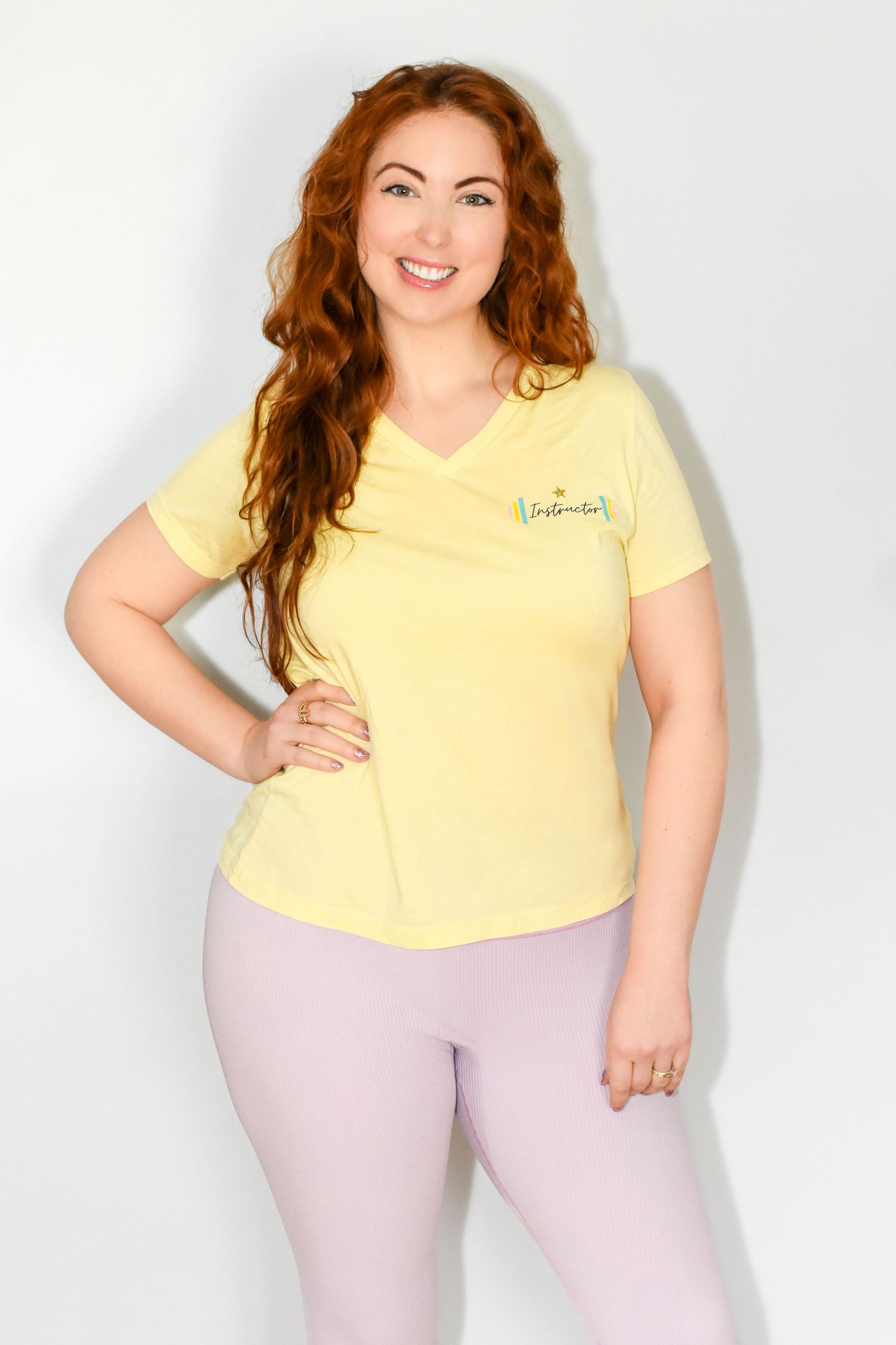 Woman wearing a yellow v-neck t-shirt and pink leggings poses confidently against a white background. Professional headshot photography.