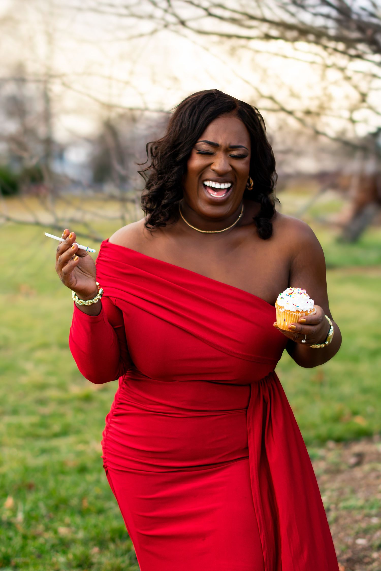 Woman in red gown laughing with cupcake and sparkler. Candid celebration portrait capturing genuine joy in park setting.