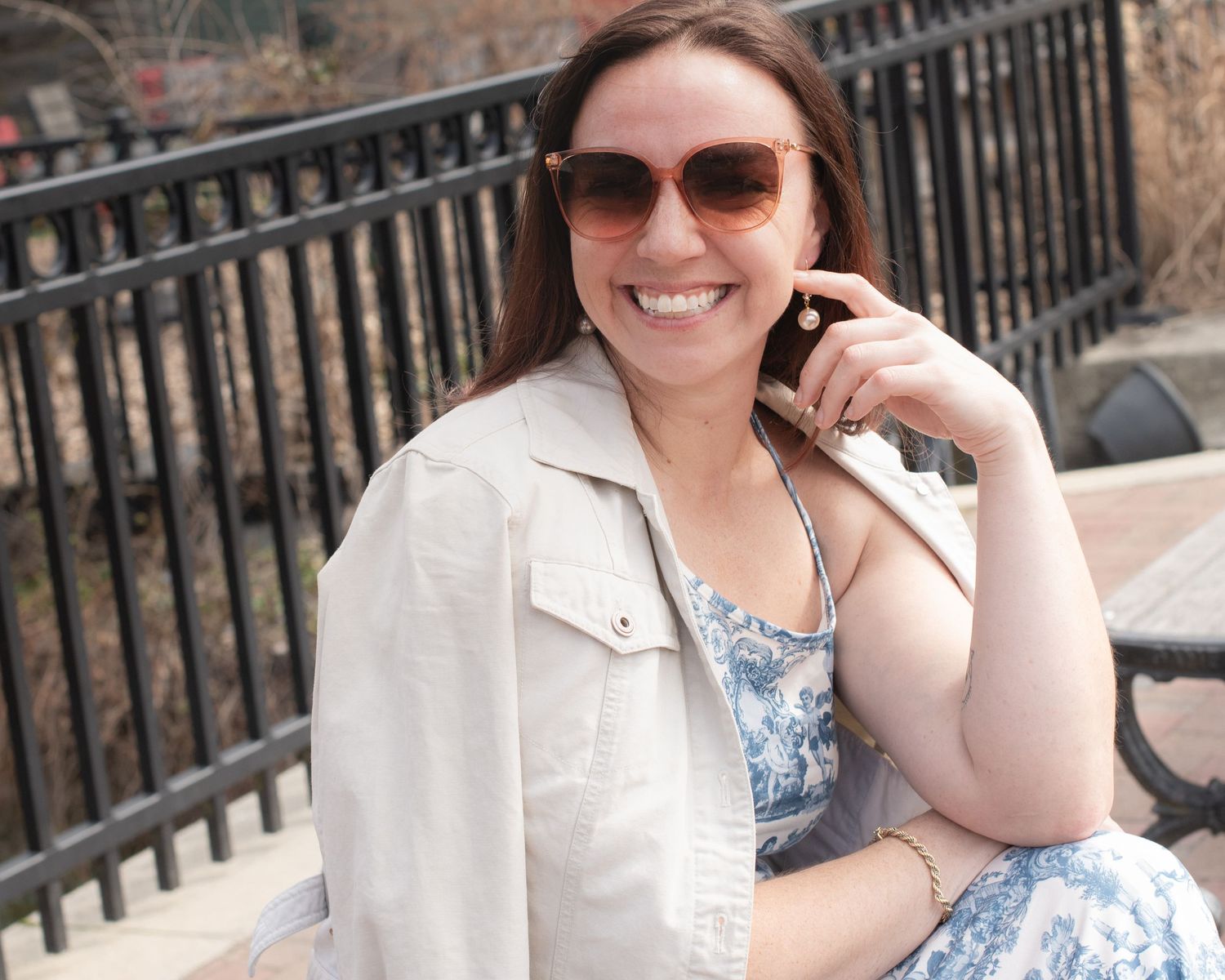 Smiling brunette woman wearing sunglasses and floral dress seated on bench outdoors in spring.
