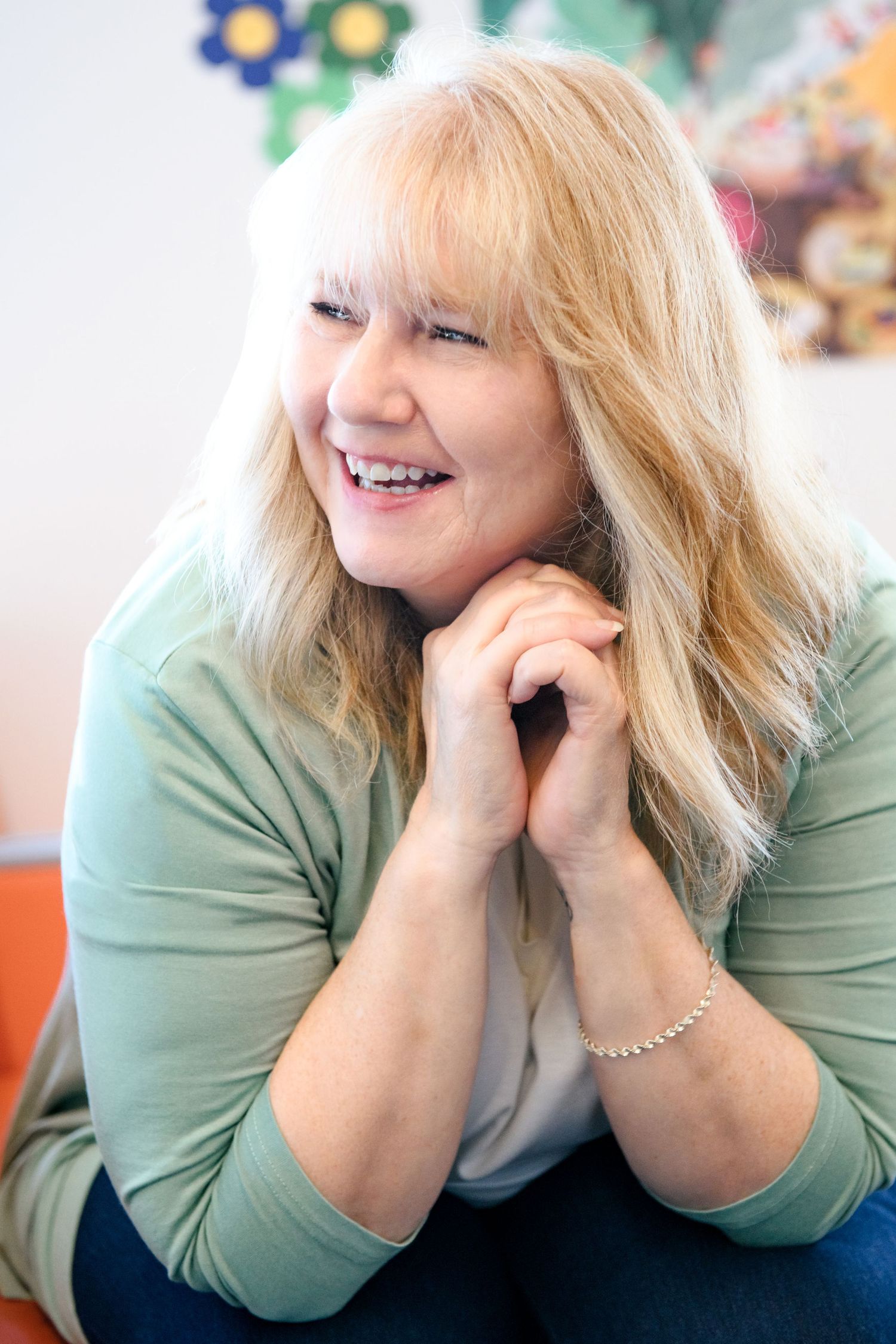 Cheerful blonde woman smiling with clasped hands in bright room with colorful wall art.