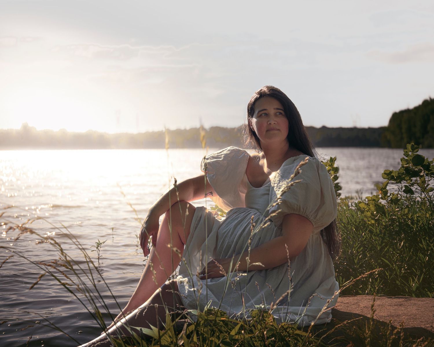 Dark-haired woman in white dress sitting by a serene lake at golden sunset surrounded by wildflowers.