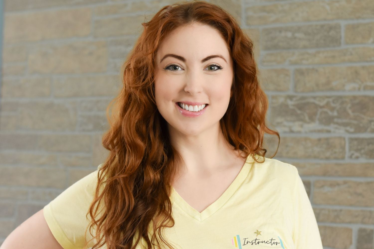 Professional headshot wearing a yellow v-neck top against a textured wall background.