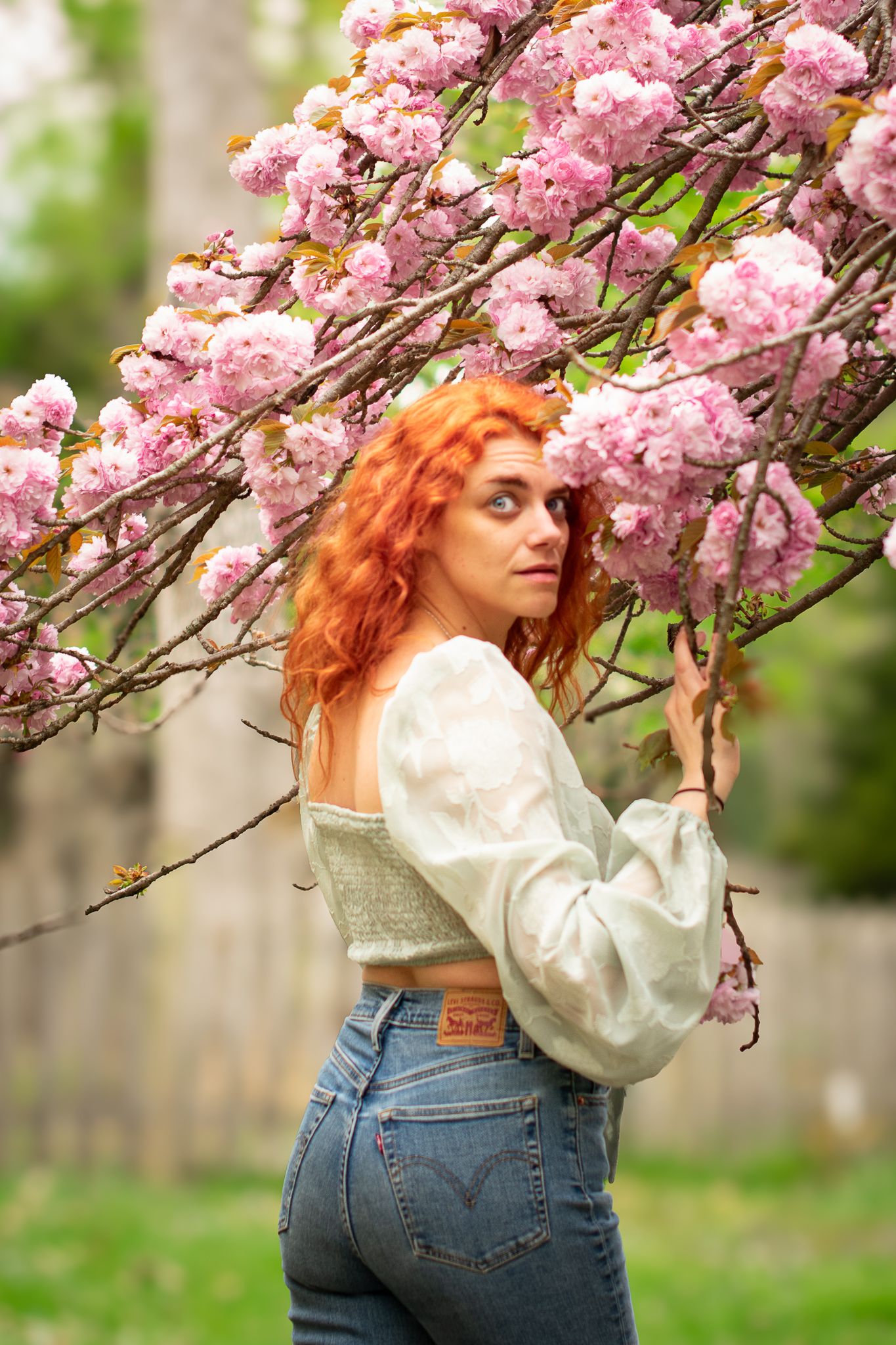 Woman wearing jeans and mint blouse stands beneath blooming pink cherry blossom branches in spring garden. Professional portrait photography