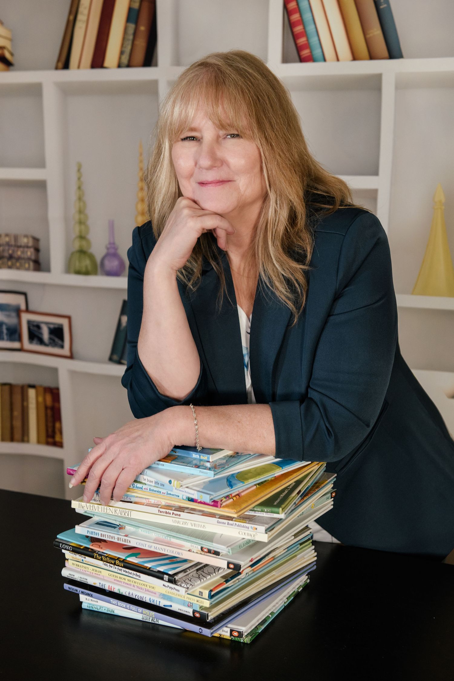 Woman in teal blazer posing with stack of colorful books, bookshelf in background.
