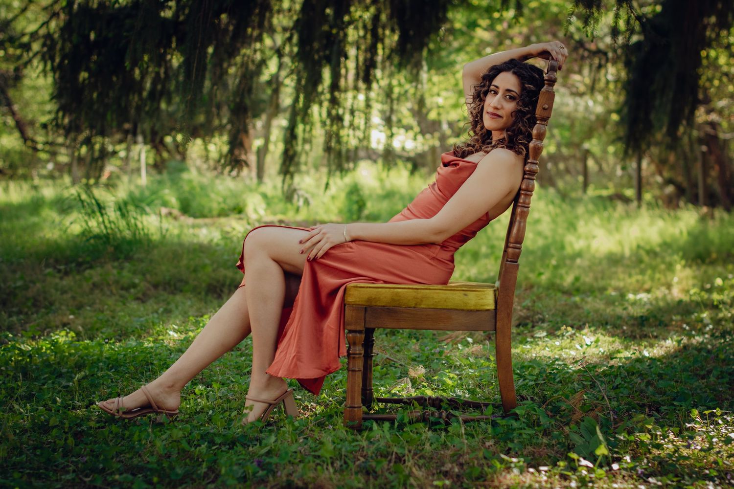 A lounging woman in a flowery dress relaxes outdoors on a wooden chair in dappled sunlight and lush greenery. Professional photography.