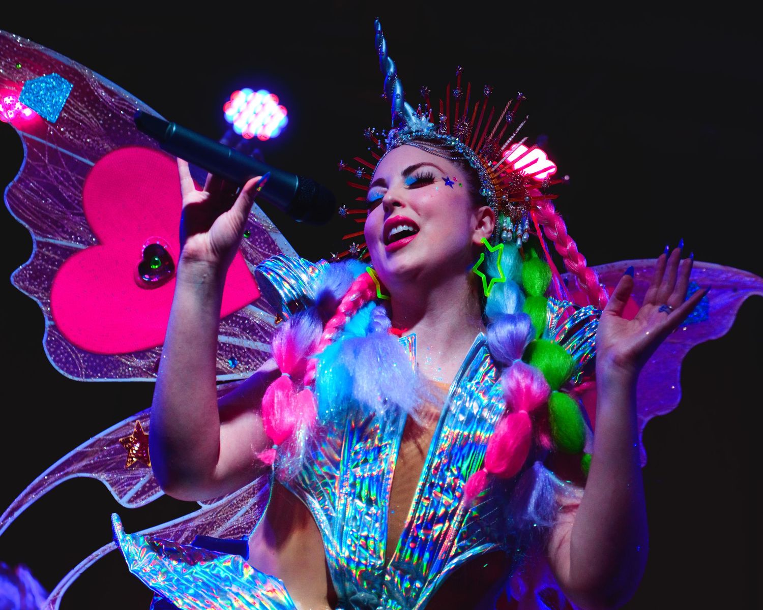 Colorful fantasy performer singing on stage wearing butterfly wings and unicorn crown under vibrant lights.