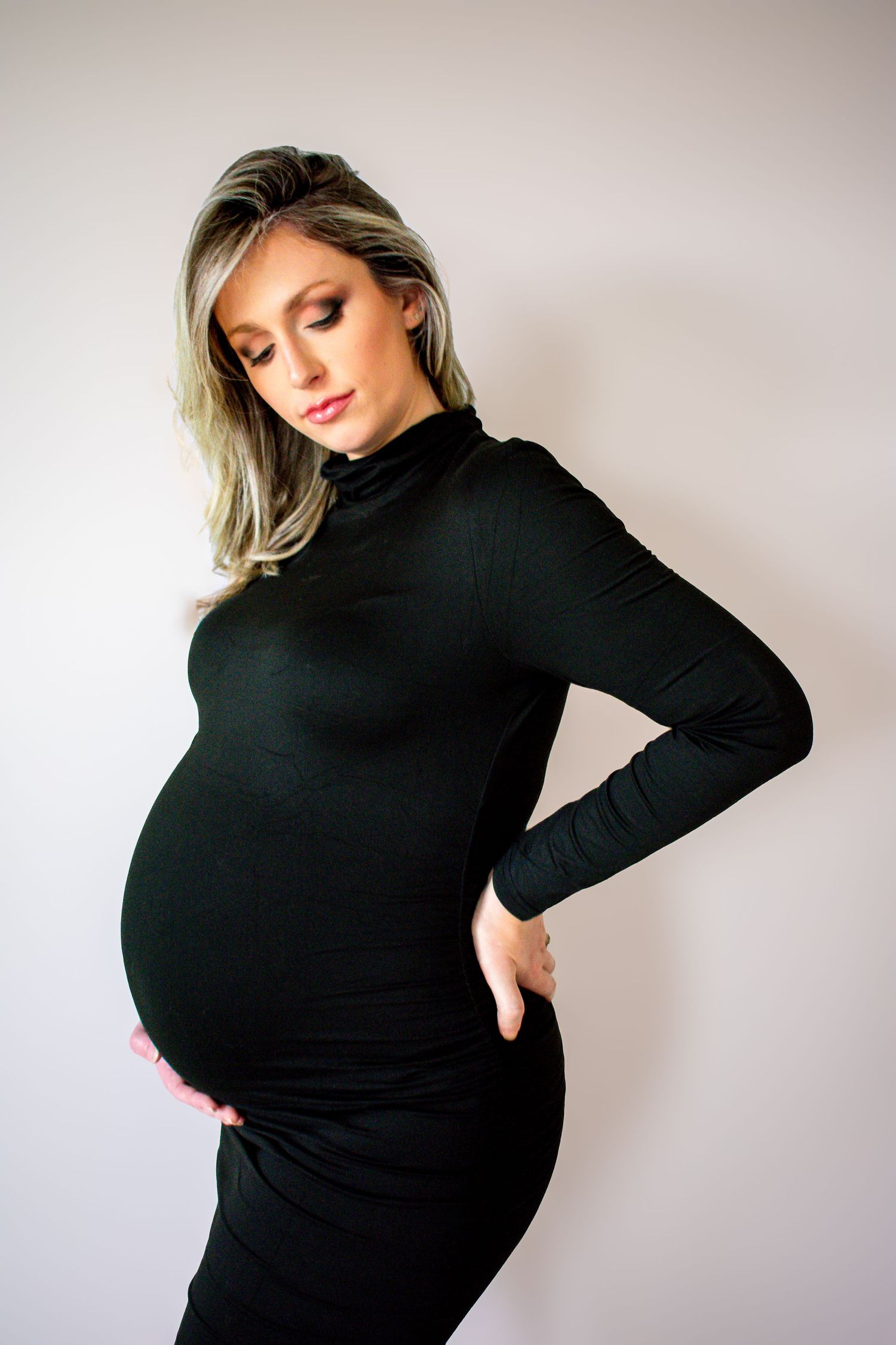 Side profile of a pregnant woman in a black turtleneck dress against a light-colored wall. Professional maternity photography