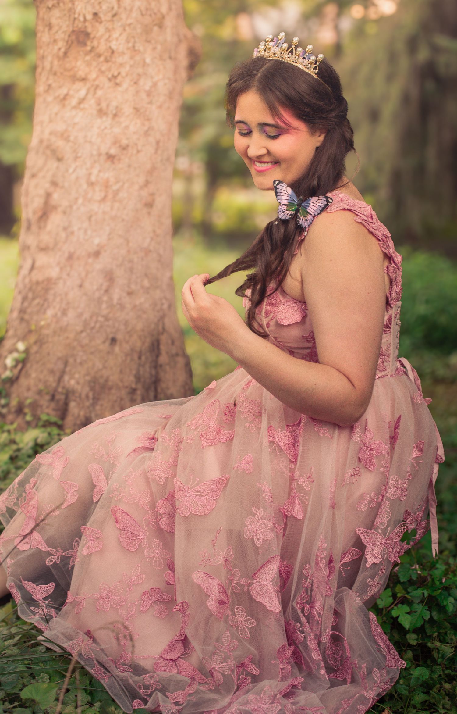 A dreamy portrait of someone in a pink floral gown and tiara sitting by a tree in a garden setting.
